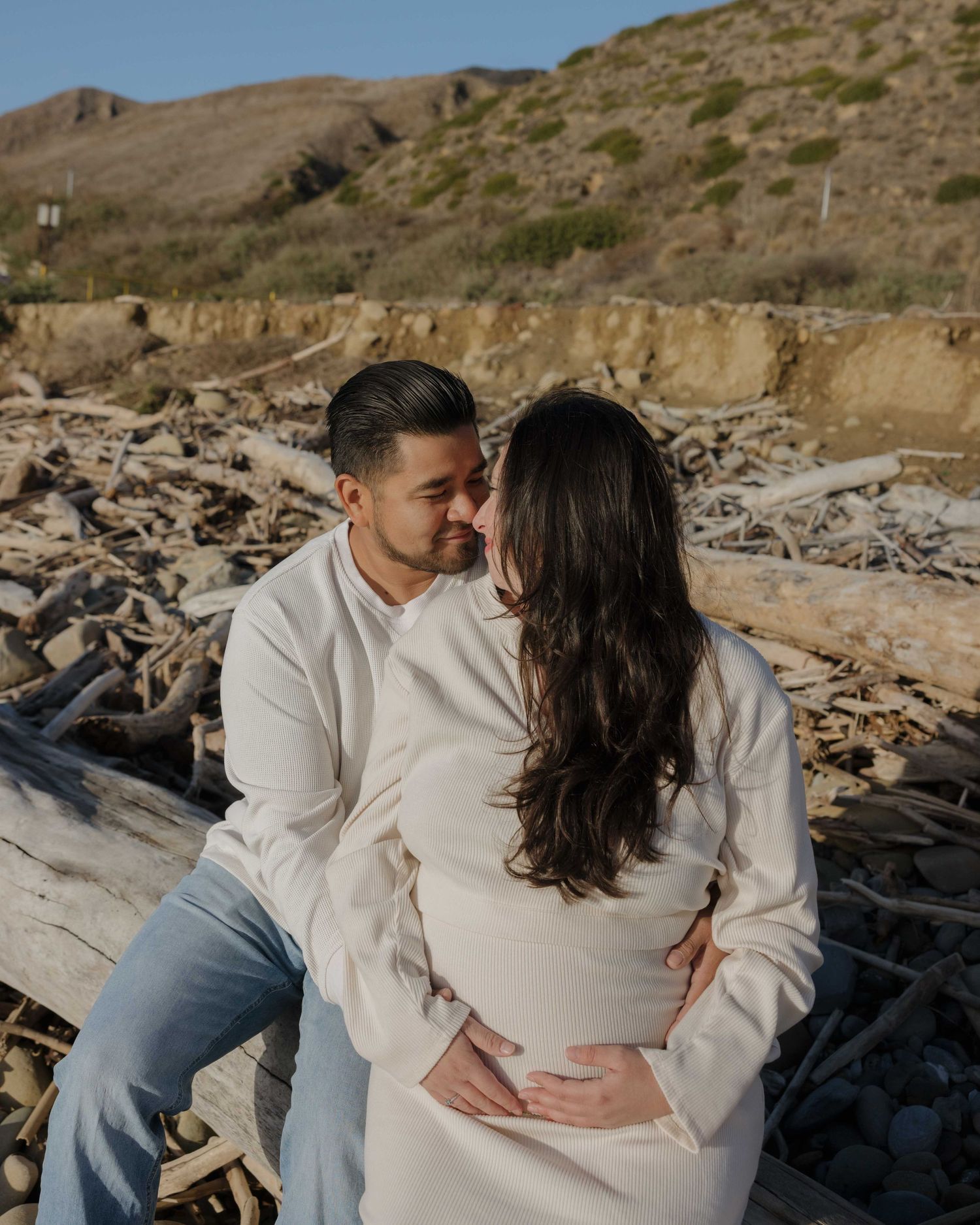 A couple in white sweaters share an intimate moment on a rocky beach with mountains in the background.