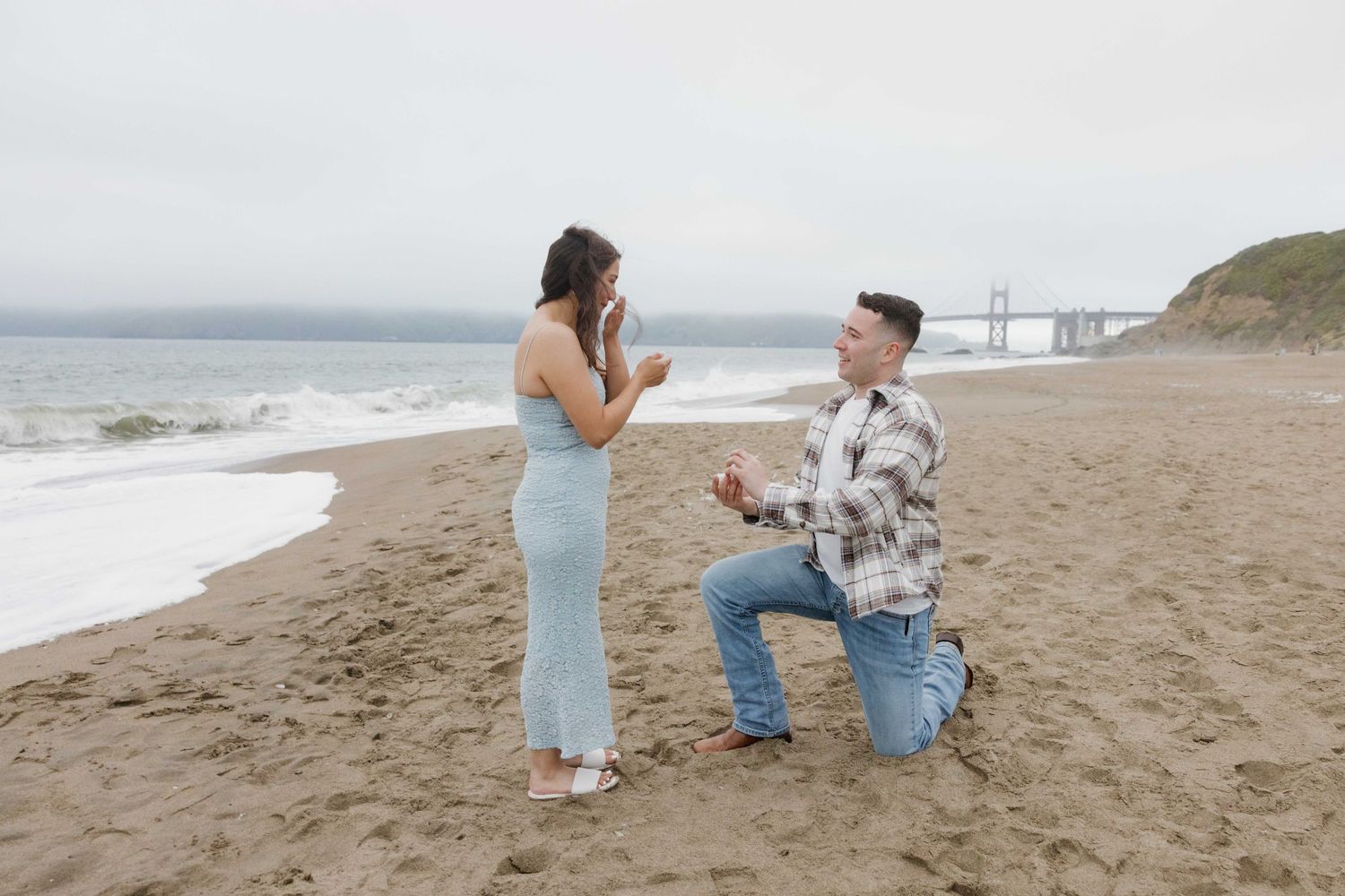 Man proposing on one knee to woman on sandy beach with Golden Gate Bridge in background.