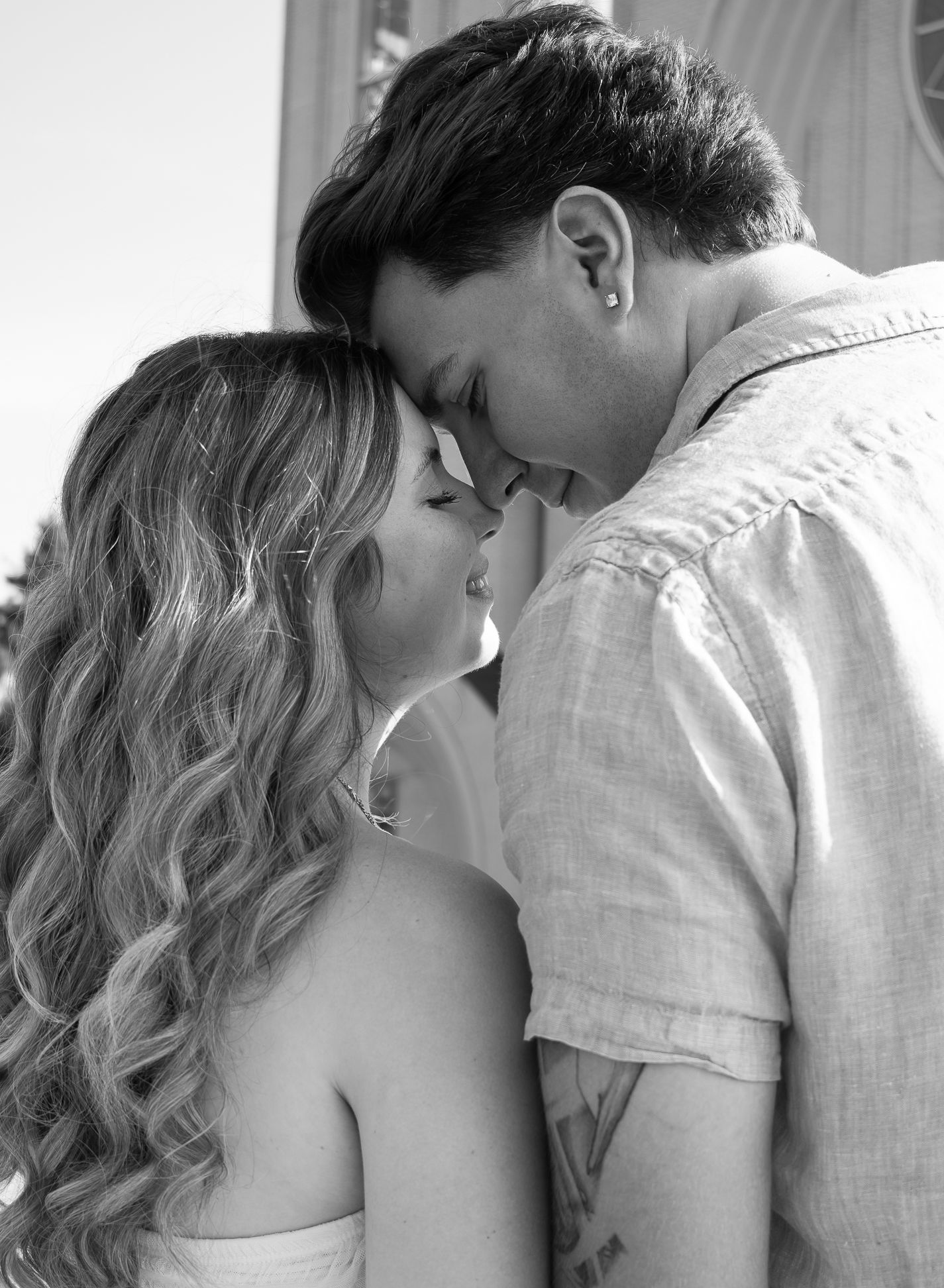 A couple standing in sunlight in front of a church, foreheads & noses touching and eyes closed while smiling, black and white image