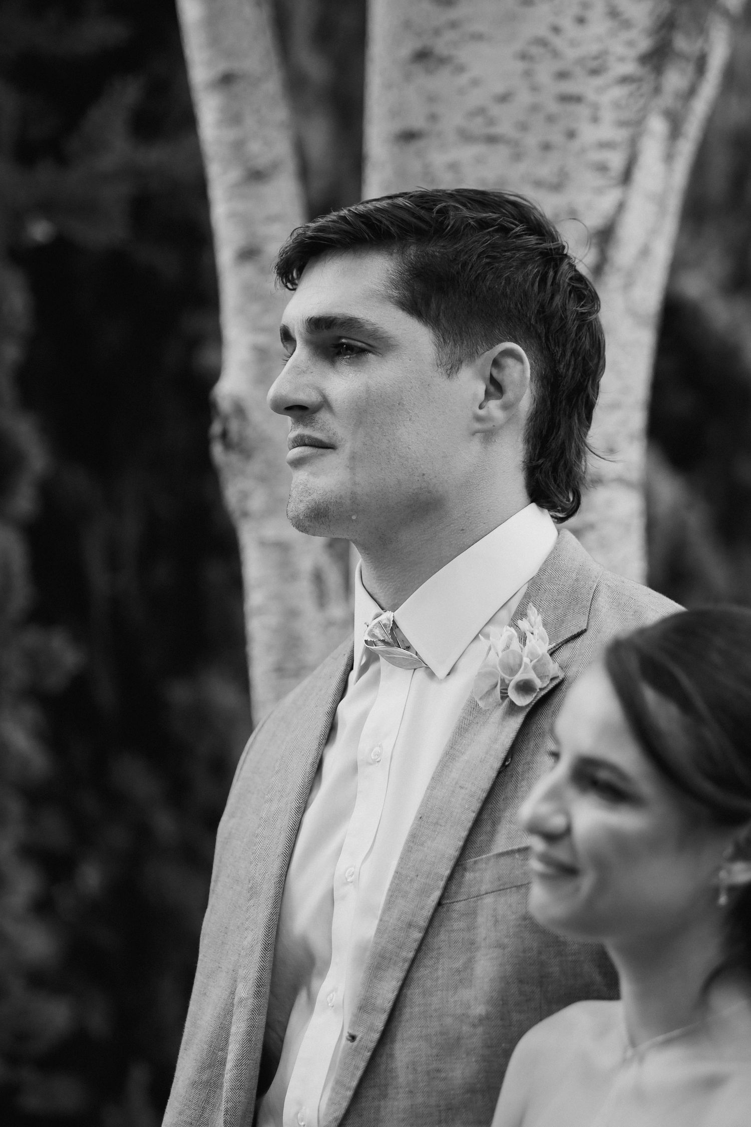 Black and white photo of a groom in a gray suit with boutonniere smiling at a wedding ceremony outdoors.