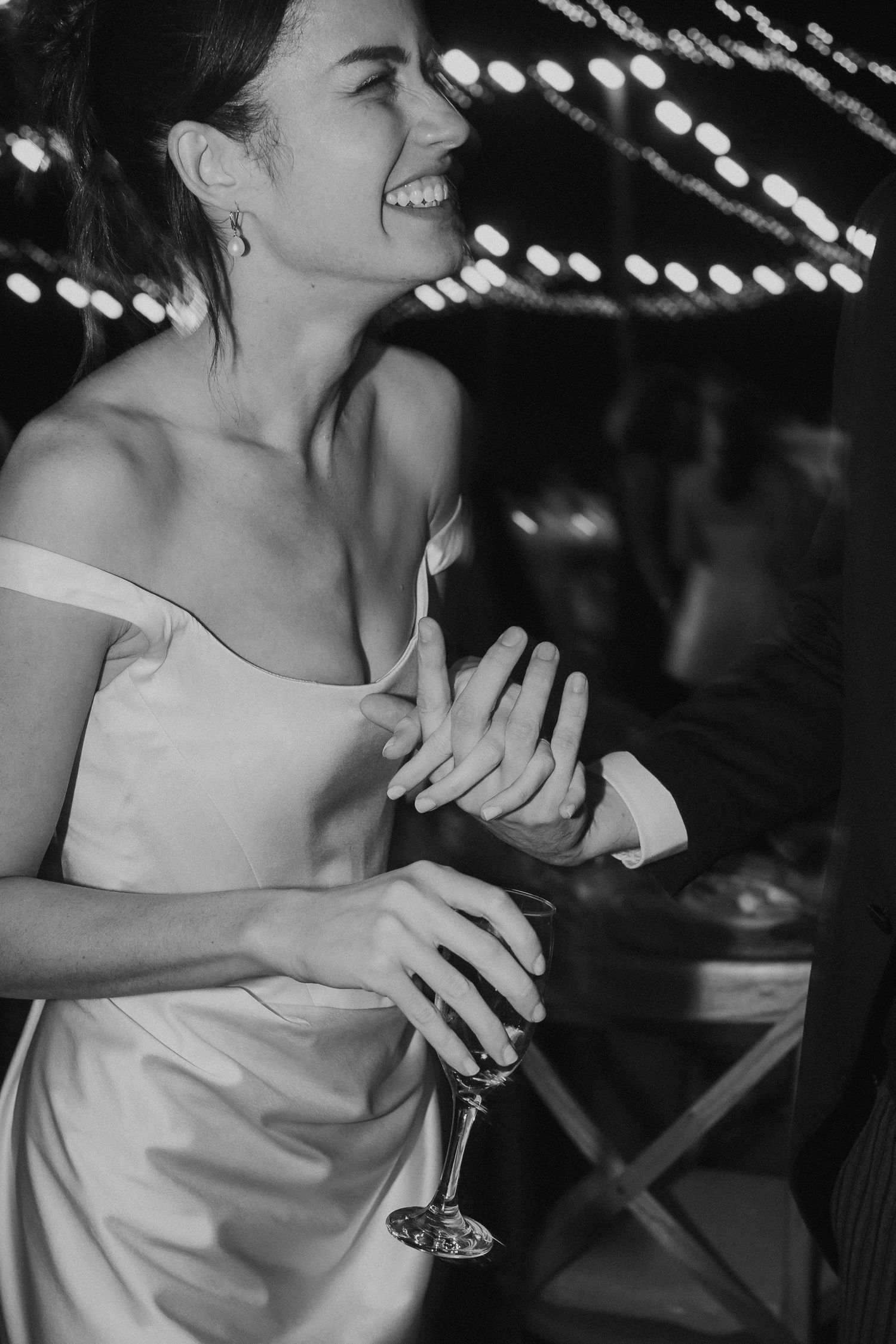 Smiling bride in an off-the-shoulder satin dress holds a wine glass and clasps hands while dancing under string lights at night.