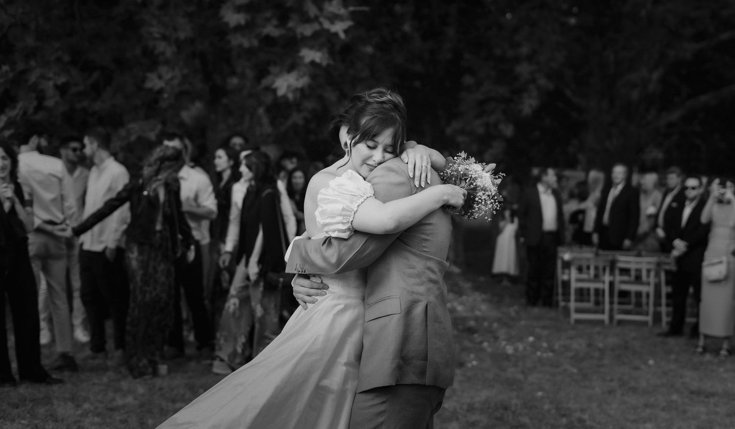 Black and white photo of a couple sharing their first dance at an outdoor wedding surrounded by guests.