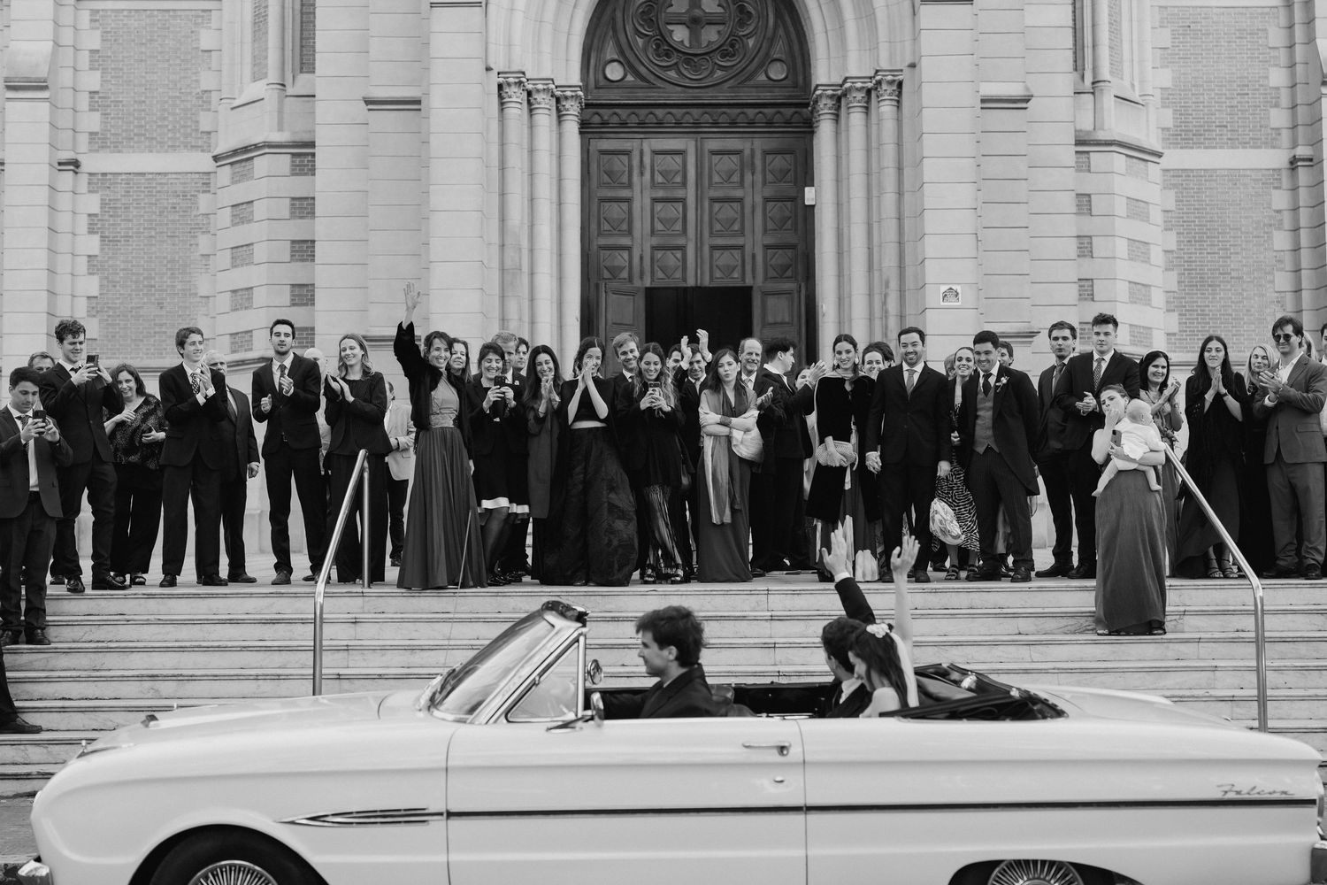 Black and white photo of wedding guests in formal attire cheering on church steps as a couple departs in a vintage white convertible.