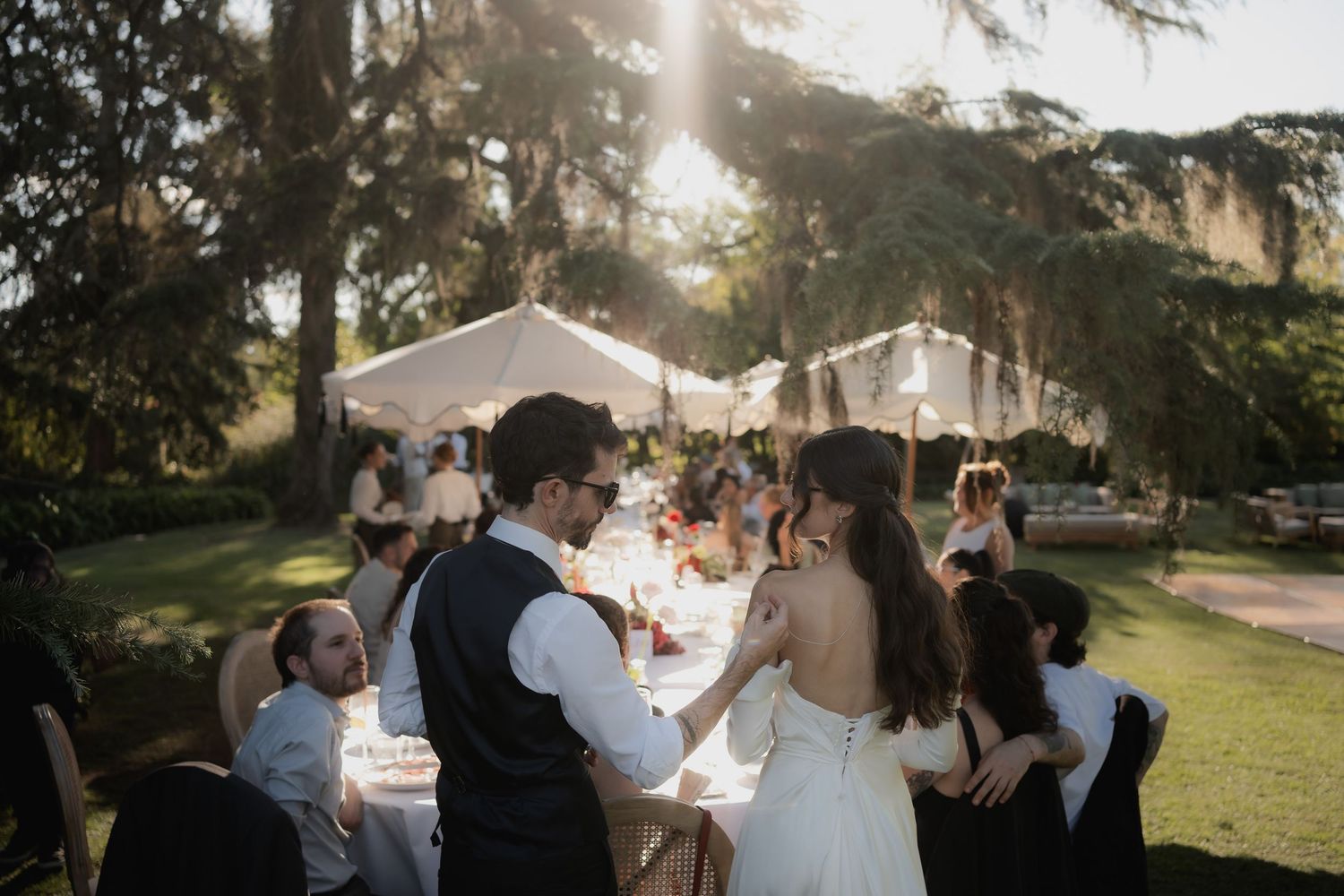 Bride and groom share a romantic moment at an elegant outdoor wedding reception under tall trees with a white tent.