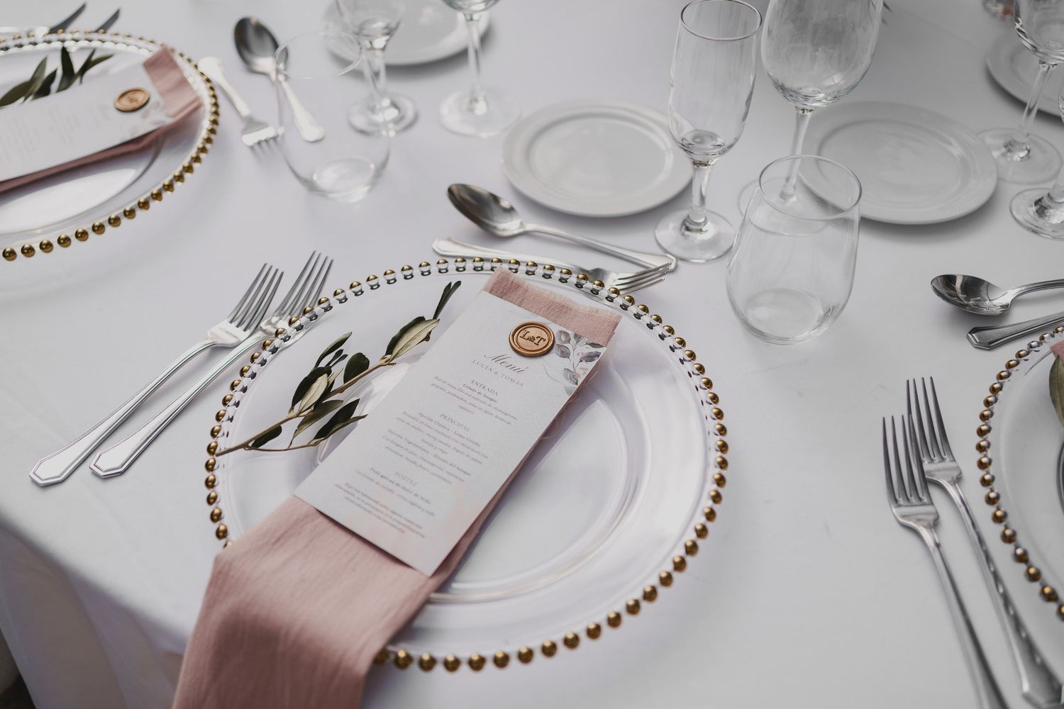 Elegant wedding table setting with gold beaded charger plates, pink napkins, silverware, and crystal glasses on white linen.