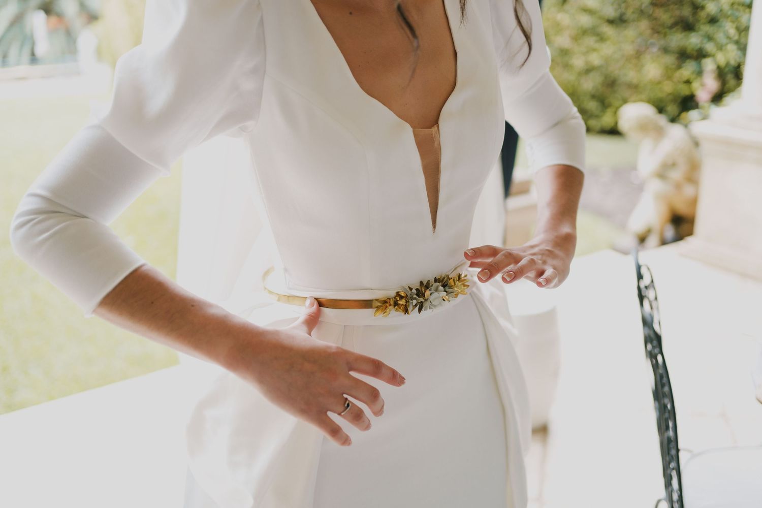 Bride in an elegant white deep V-neck wedding dress with long sleeves and a gold floral embellished belt, standing outdoors.