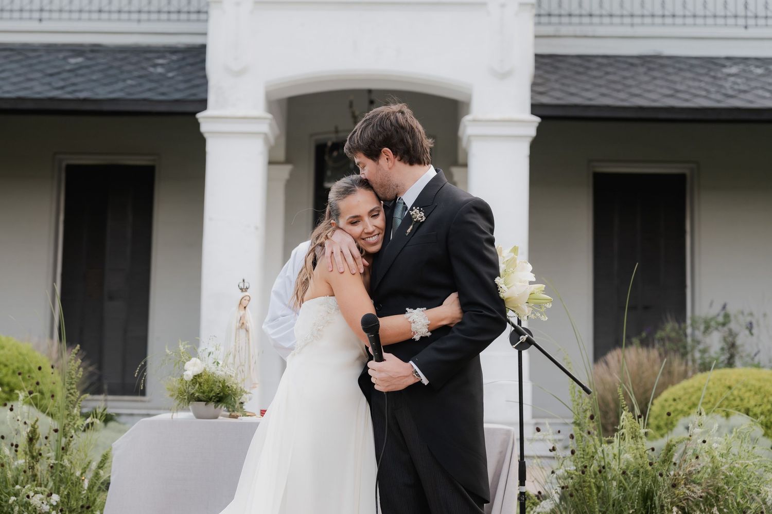 Bride in a white gown and groom in a dark suit embrace tenderly at an outdoor wedding ceremony in front of a elegant white house.