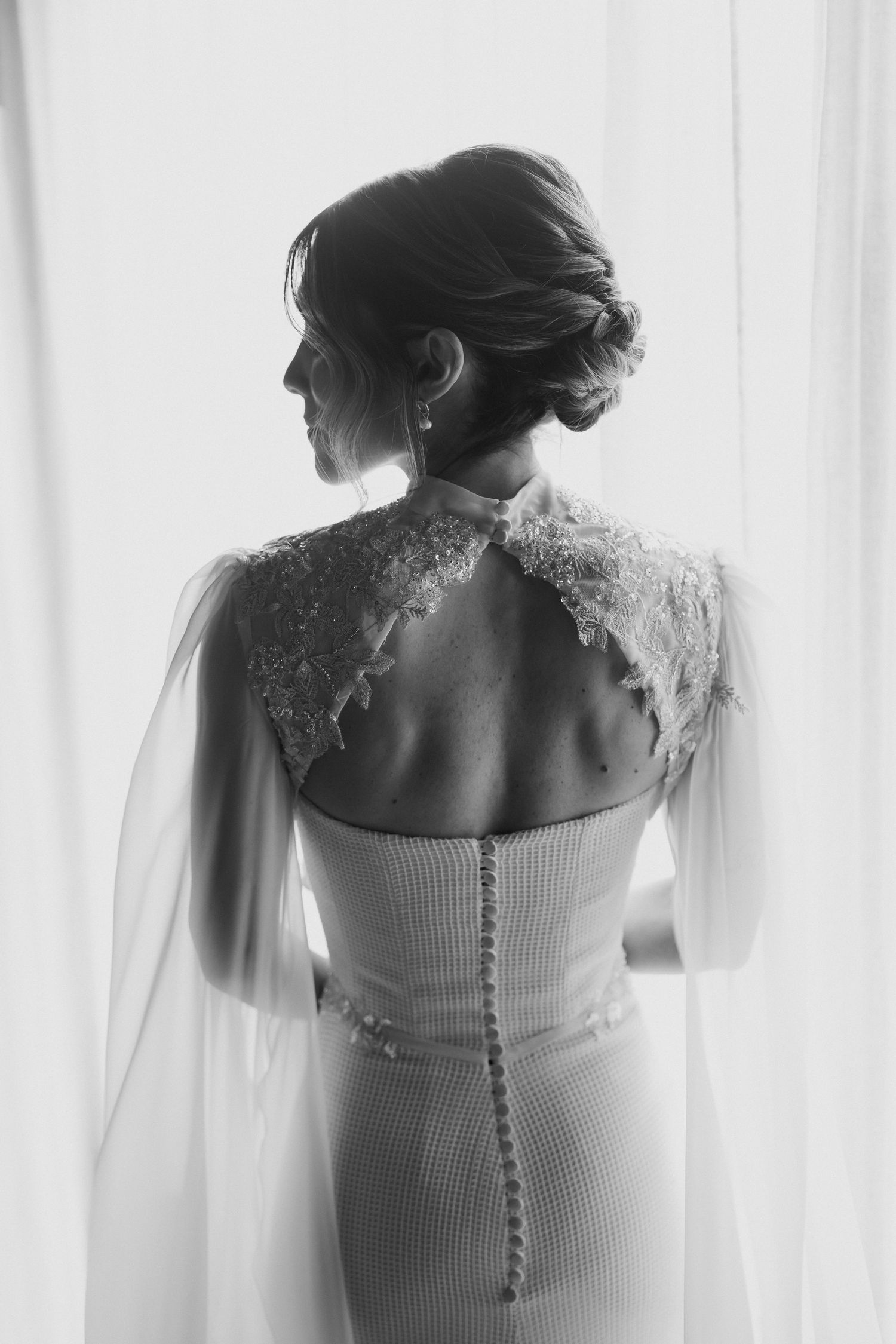 Black and white photo of a bride from behind in a lace-shouldered wedding gown with sheer cape sleeves and buttoned back.
