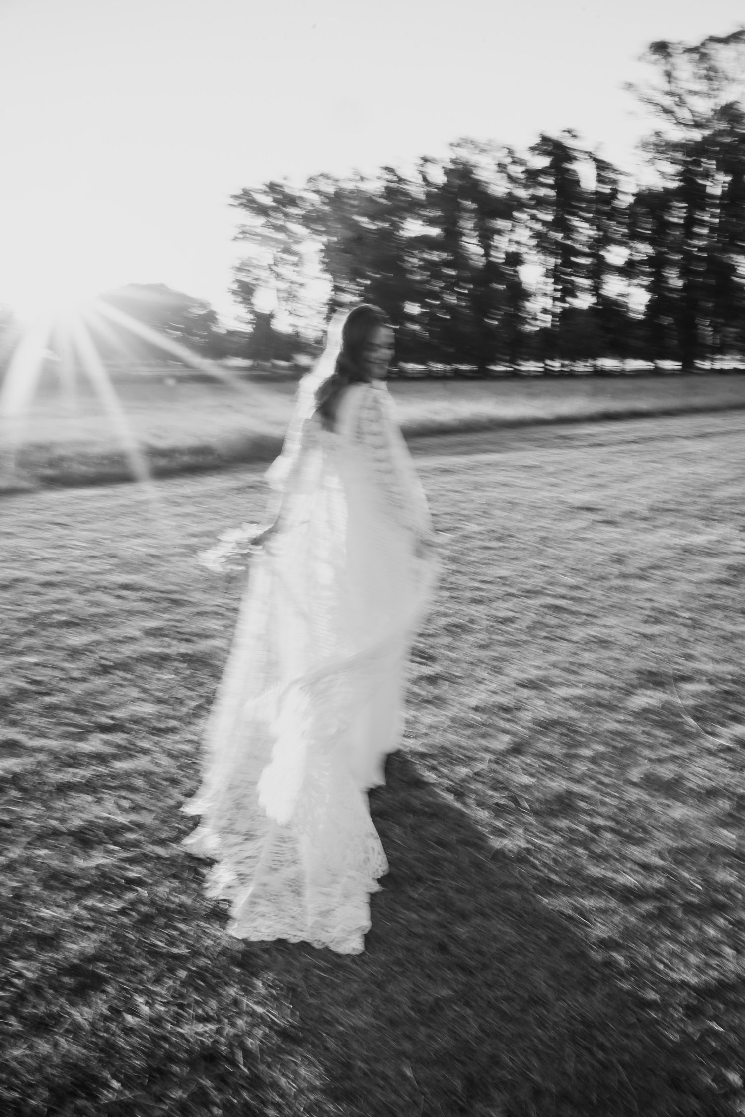 Black and white photo of a bride in a flowing gown twirling outdoors at sunset with sun flare.