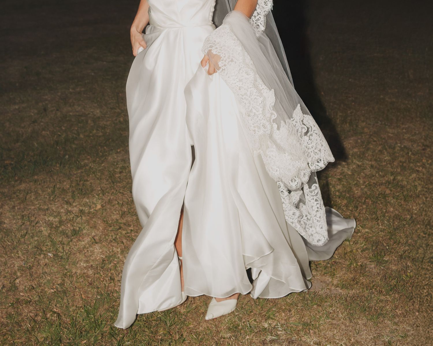 Bride wearing elegant white satin wedding gown with lace veil at night outdoor ceremony.