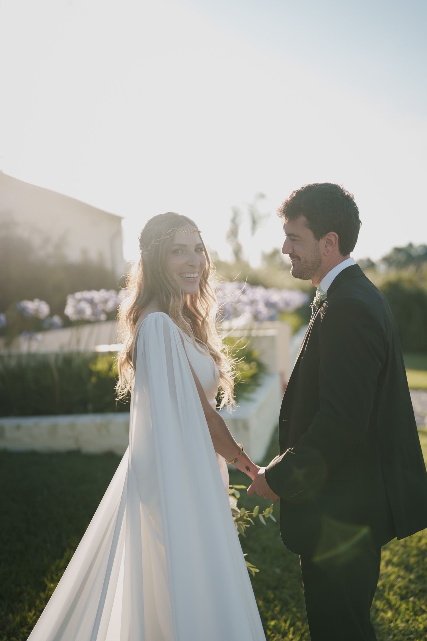 Bride in white cape gown holds hands with groom in dark suit, smiling outdoors in golden sunlight.