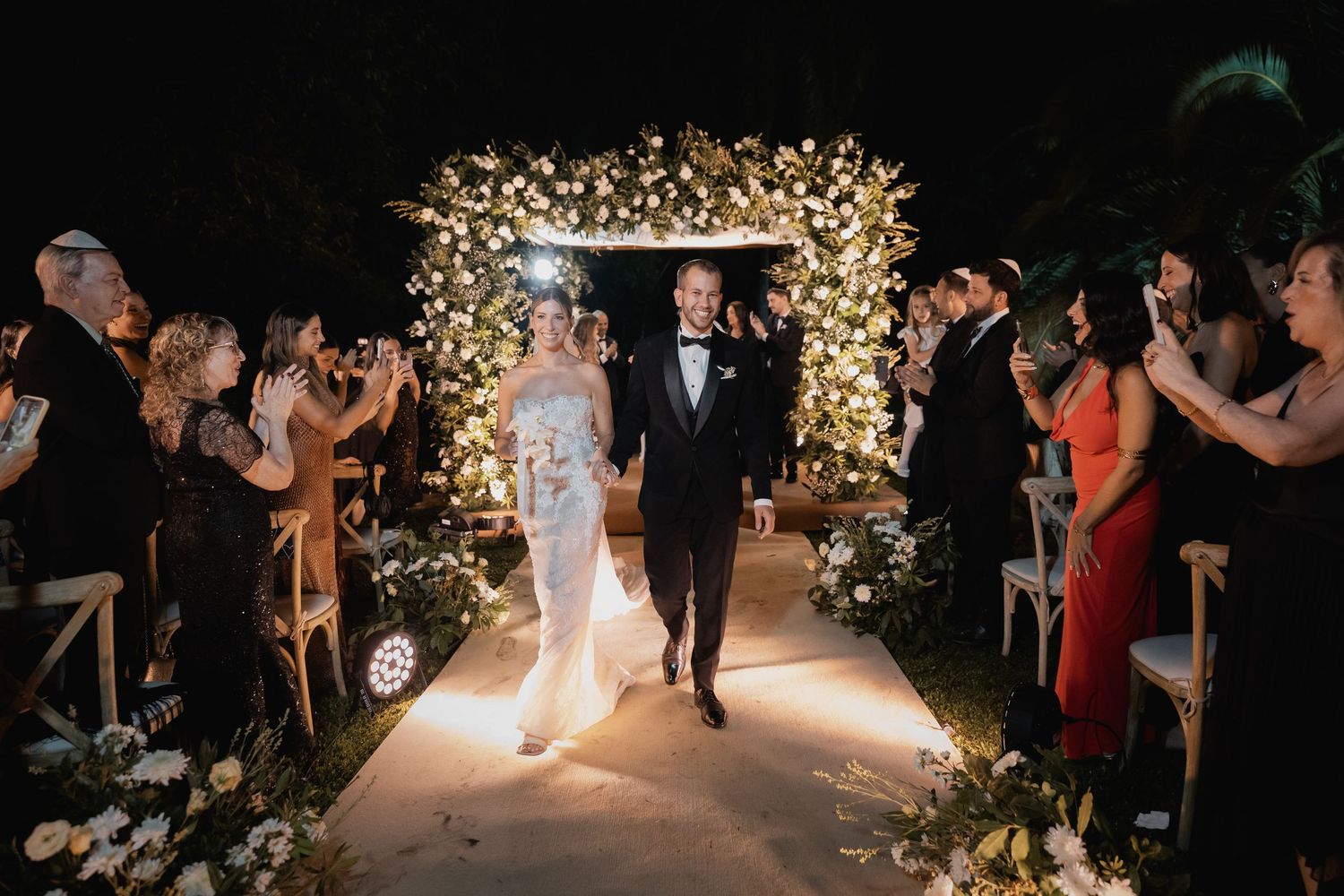 Bride and groom walk down floral arch-lit aisle at elegant outdoor night wedding ceremony with guests applauding.