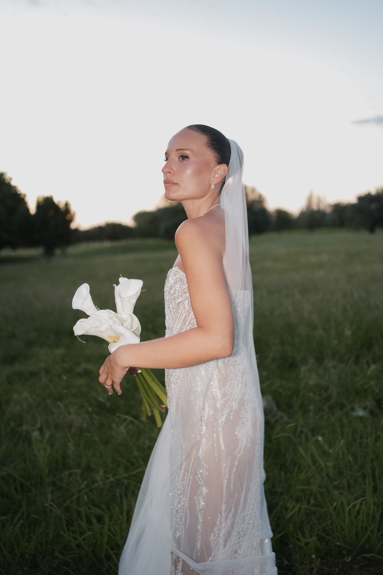 Elegant bride in white lace gown holding calla lilies, wearing veil, standing in green field at dusk.