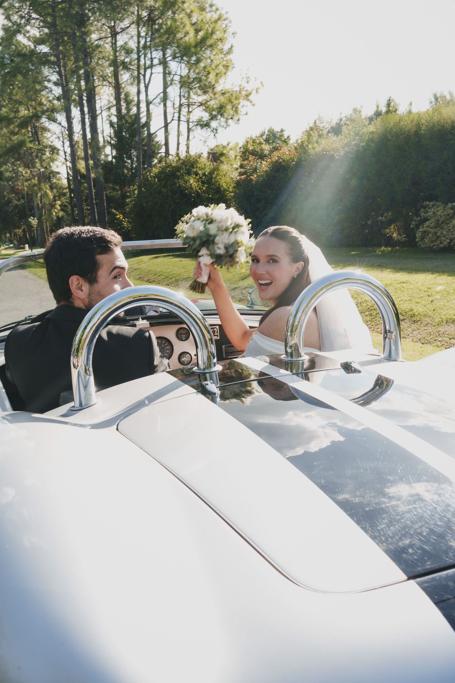 Joyful bride holding bouquet aloft in classic white convertible sports car with groom at sunny outdoor venue.