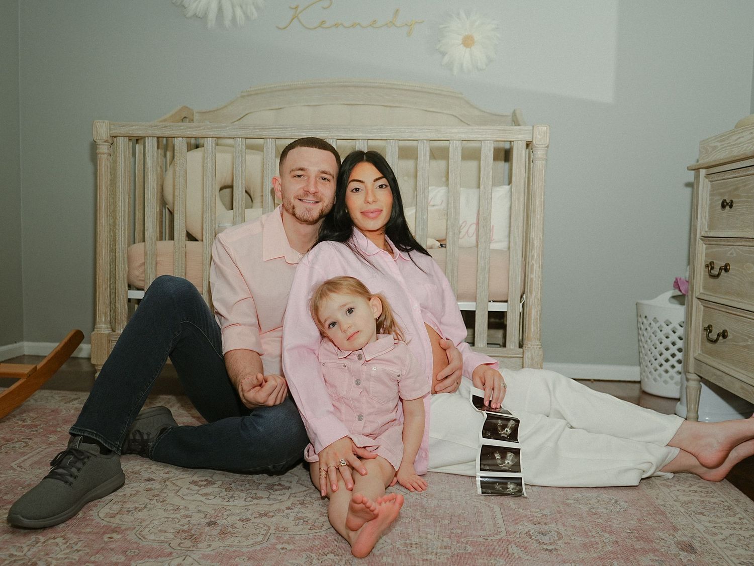Three people sit together on the floor of a nursery room holding ultrasound photos and wearing matching pink outfits.