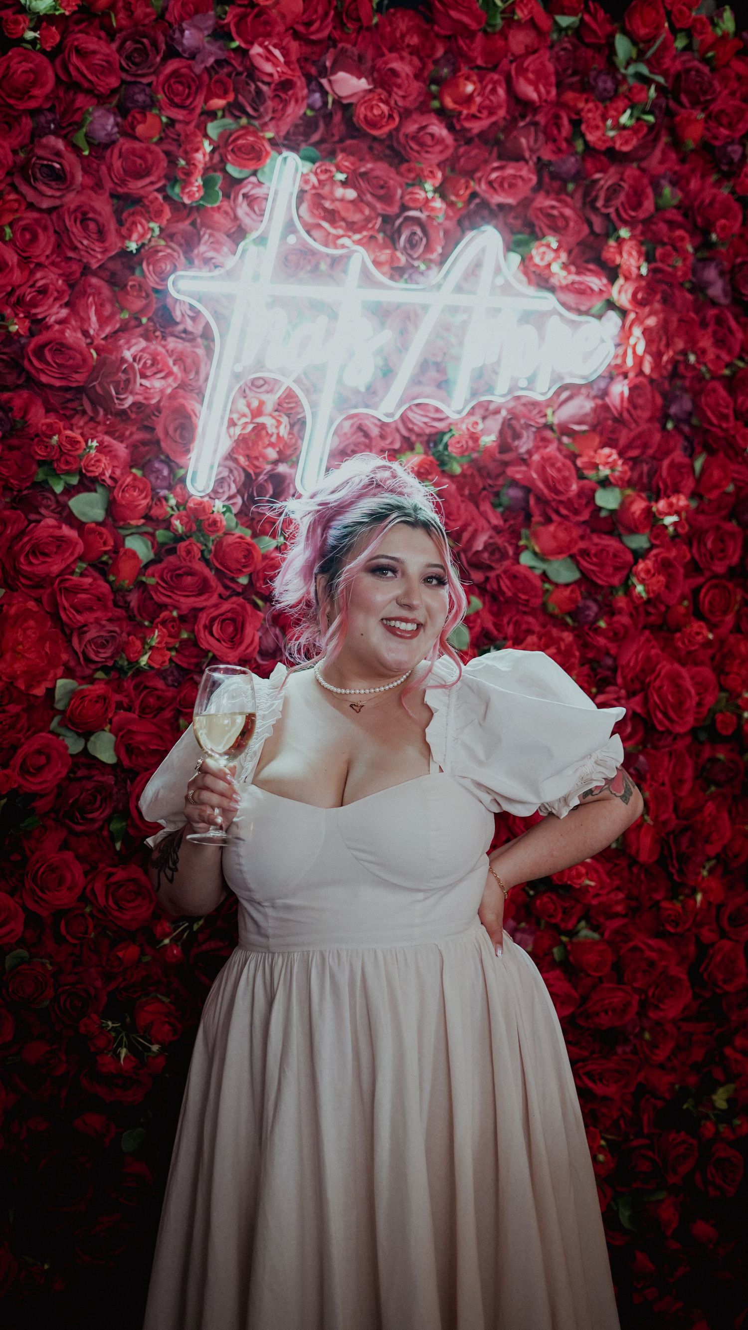 A person in a flowing ivory dress poses with champagne against a dramatic red rose wall with a neon light sign.