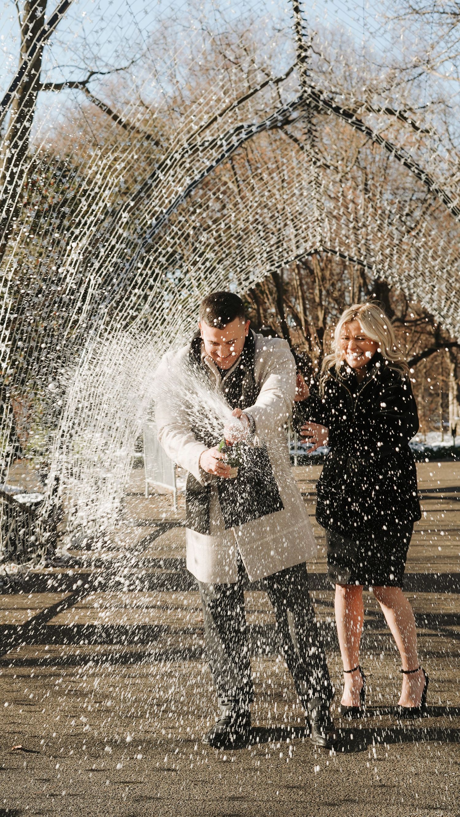 A couple playfully splashes through water sprinklers in a park on a sunny day.