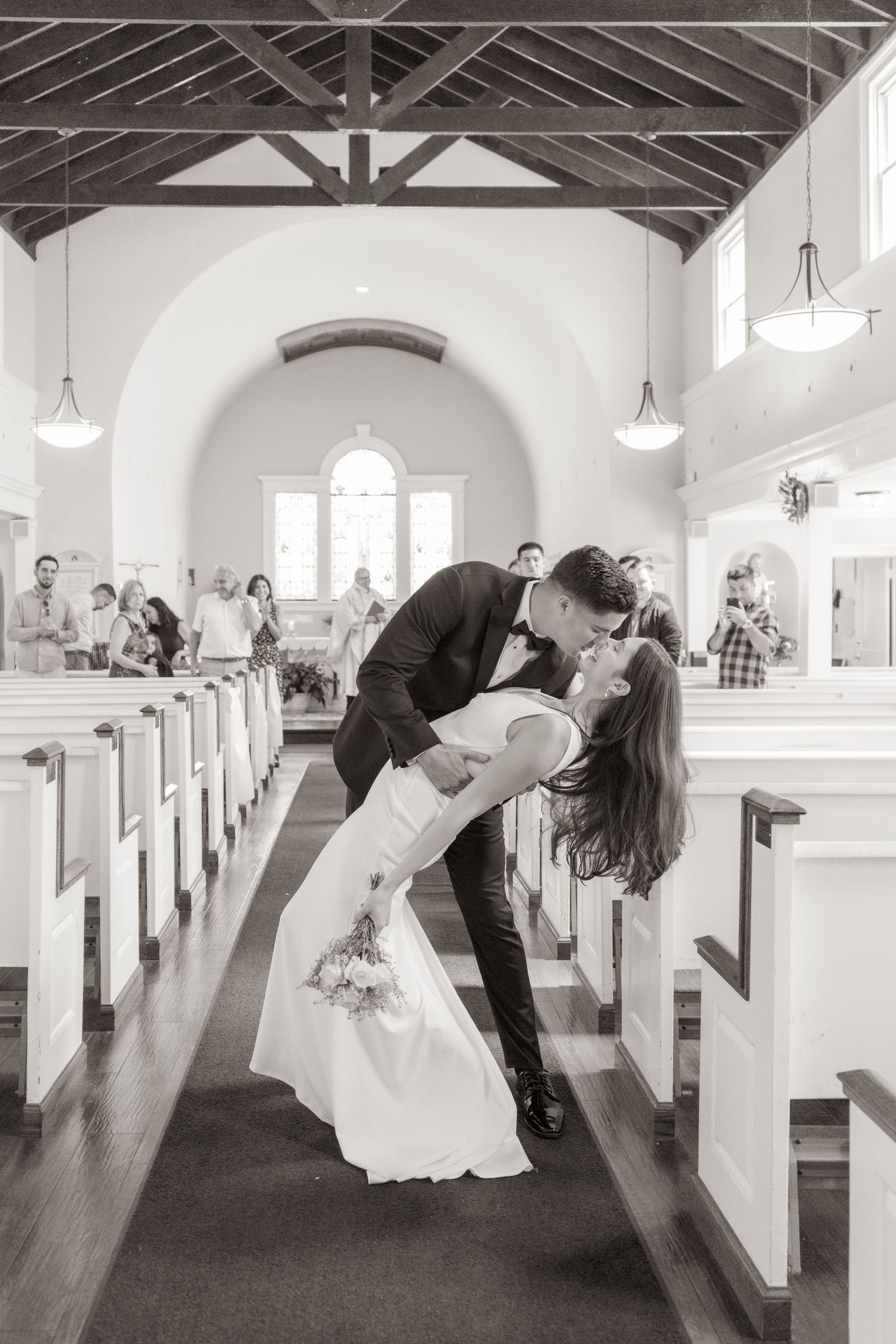 Couple shares romantic kiss in church aisle surrounded by white pews and arched ceiling in black and white photo.