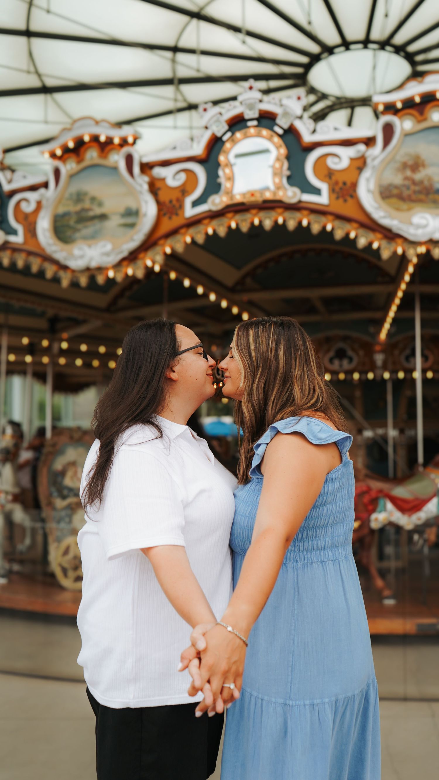 A couple shares a tender moment while holding hands in front of a vintage carousel with ornate decorative details.