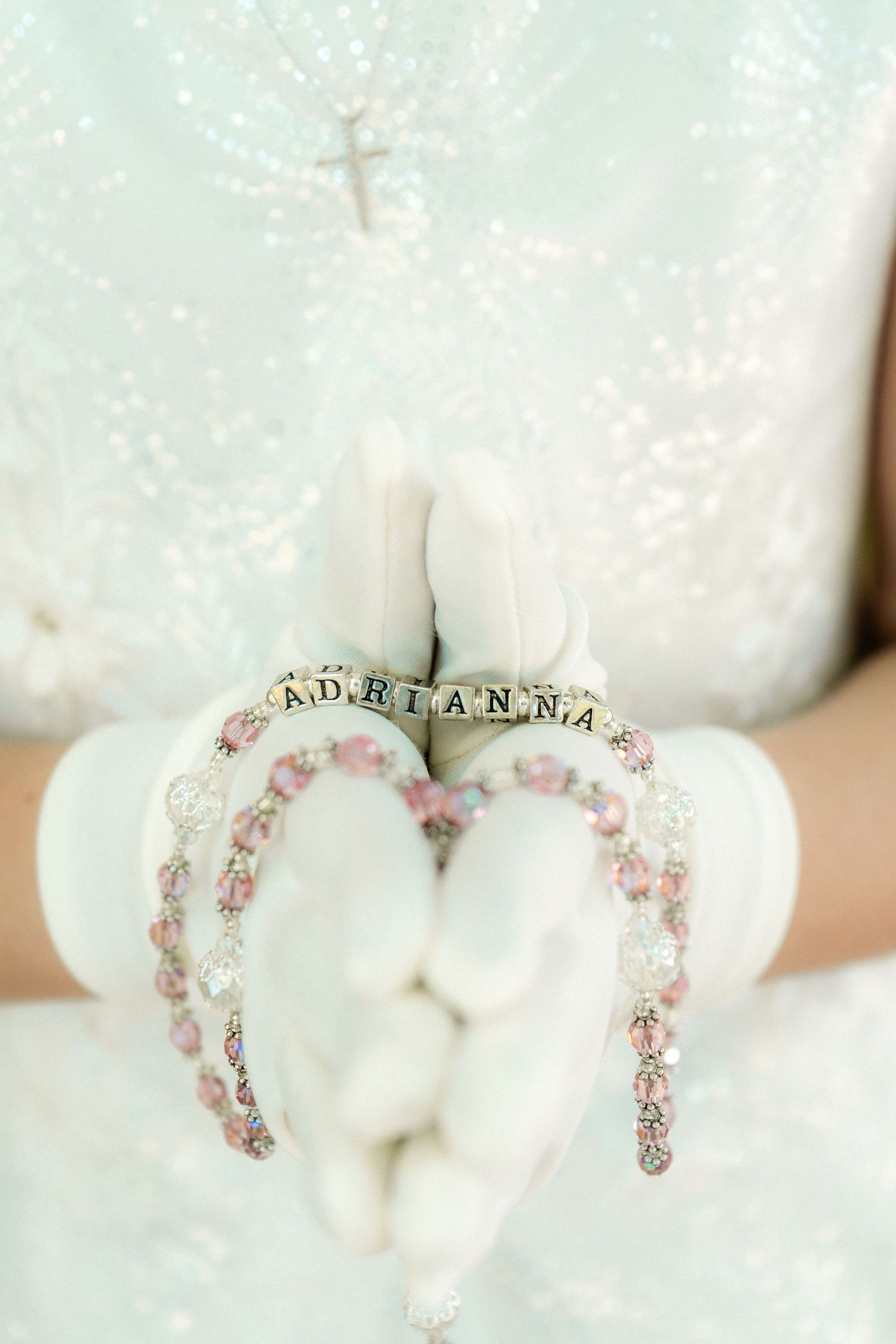 A delicate beaded jewelry piece spelling 'Adrianna' in pink and clear crystals against a soft white background.