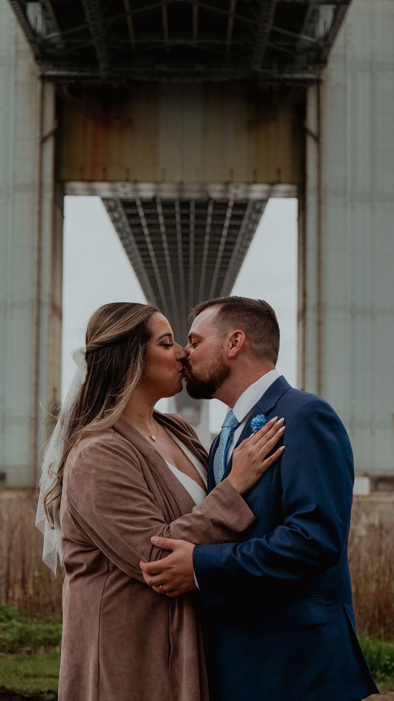 A romantic couple shares a tender kiss under a bridge structure while dressed in formal attire.