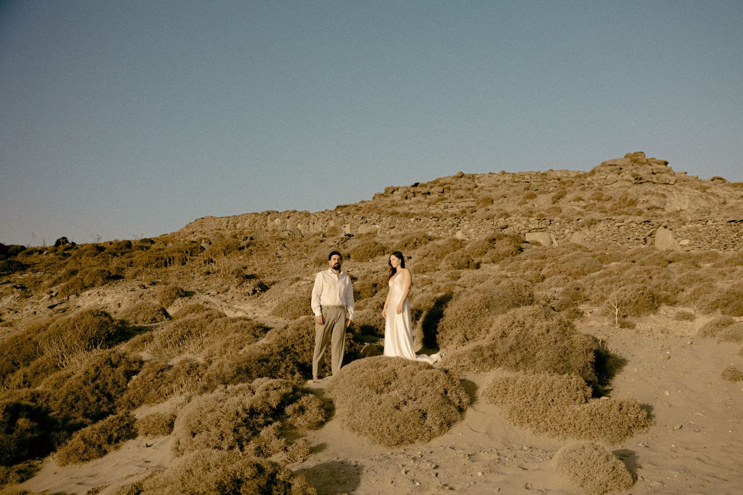 A couple in wedding attire poses on rocky terrain during golden hour. Wedding photographer in Kythnos.