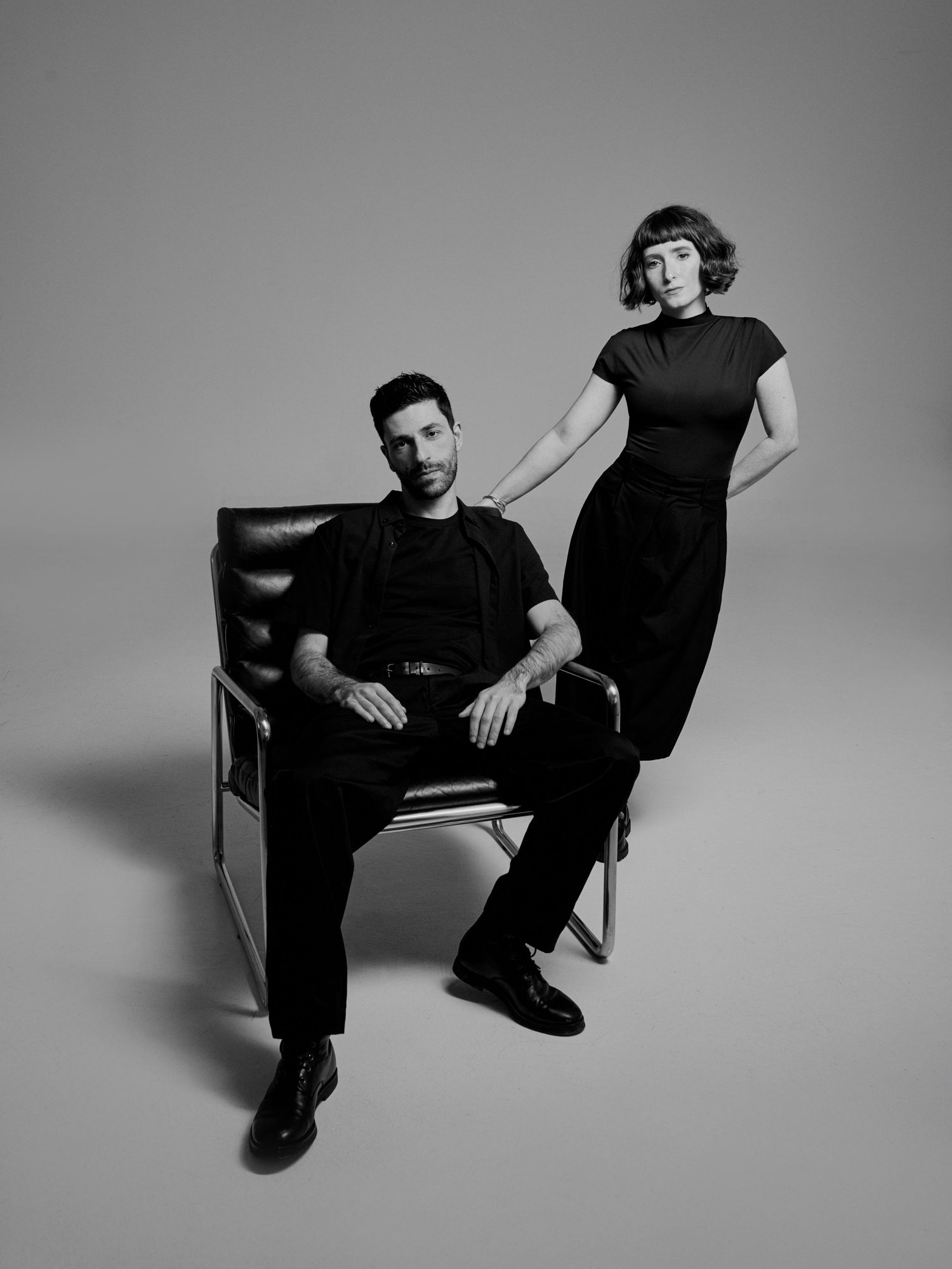 Black and white portrait of a man seated in a chair and a woman standing beside him, both dressed in elegant black outfits.