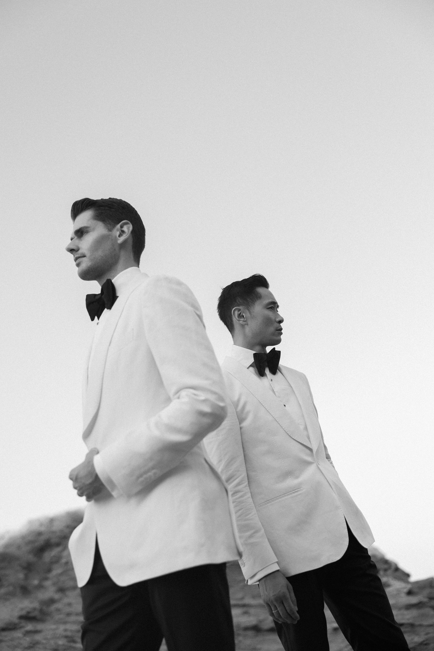 Same sex couple destination wedding LGBTQ+ photographer and bow ties stand back-to-back outdoors in a black and white photo.