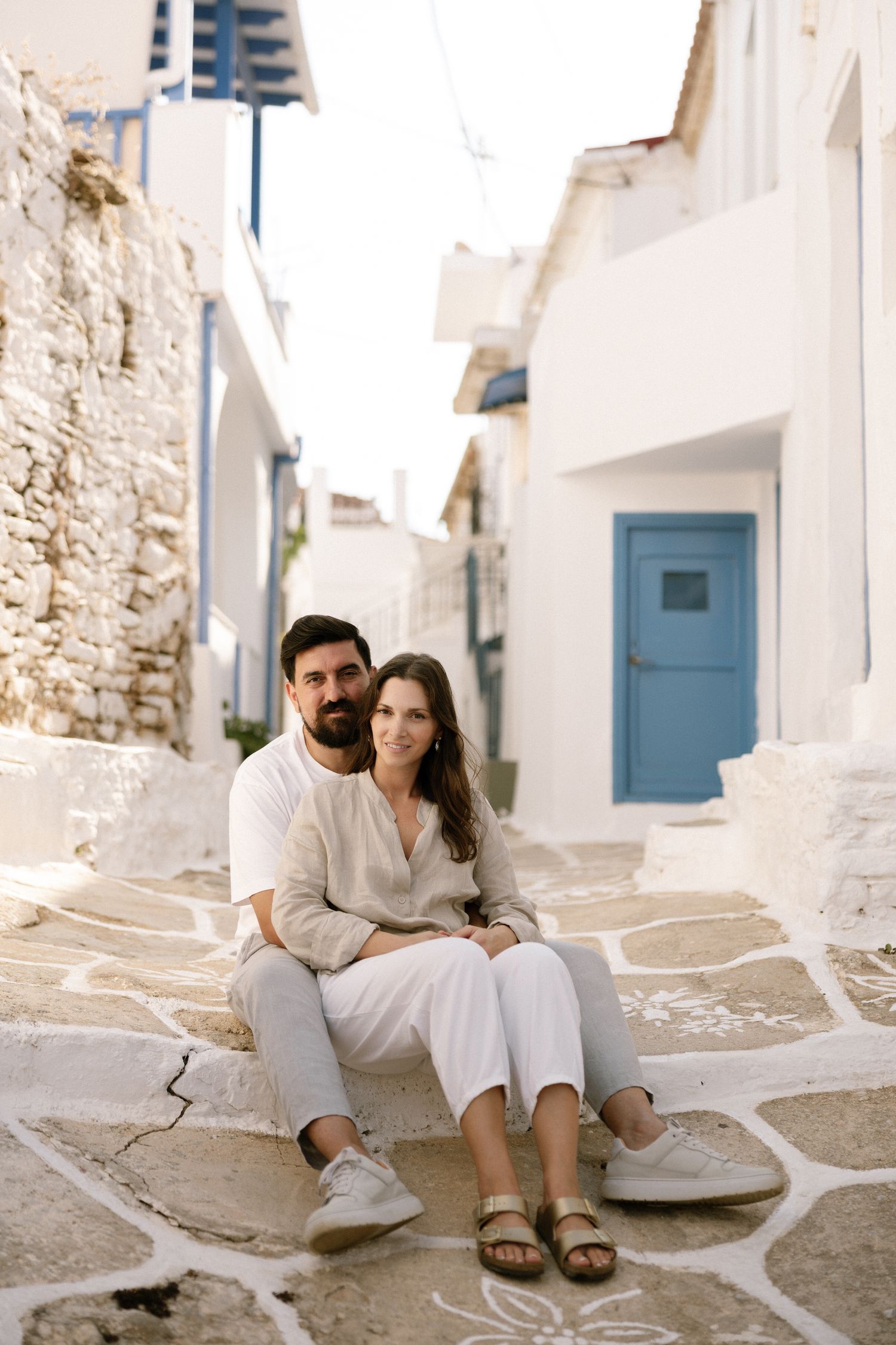 A couple in white clothing sitting on traditional Mediterranean stone steps with a blue door in Kythnos island.