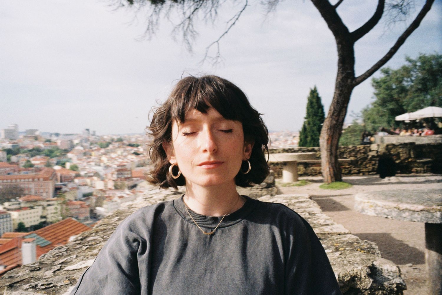 Person with short dark hair sitting outdoors overlooking a Mediterranean city landscape.