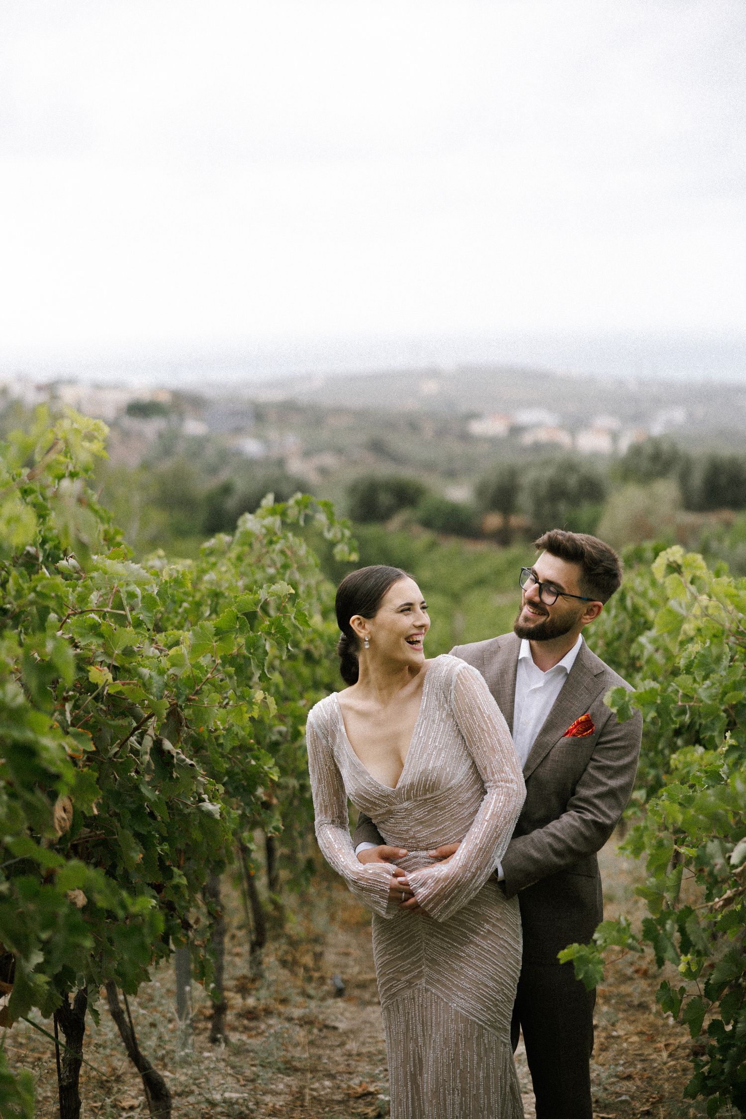 A couple laughing together in a hilltop vineyard wearing elegant attire.