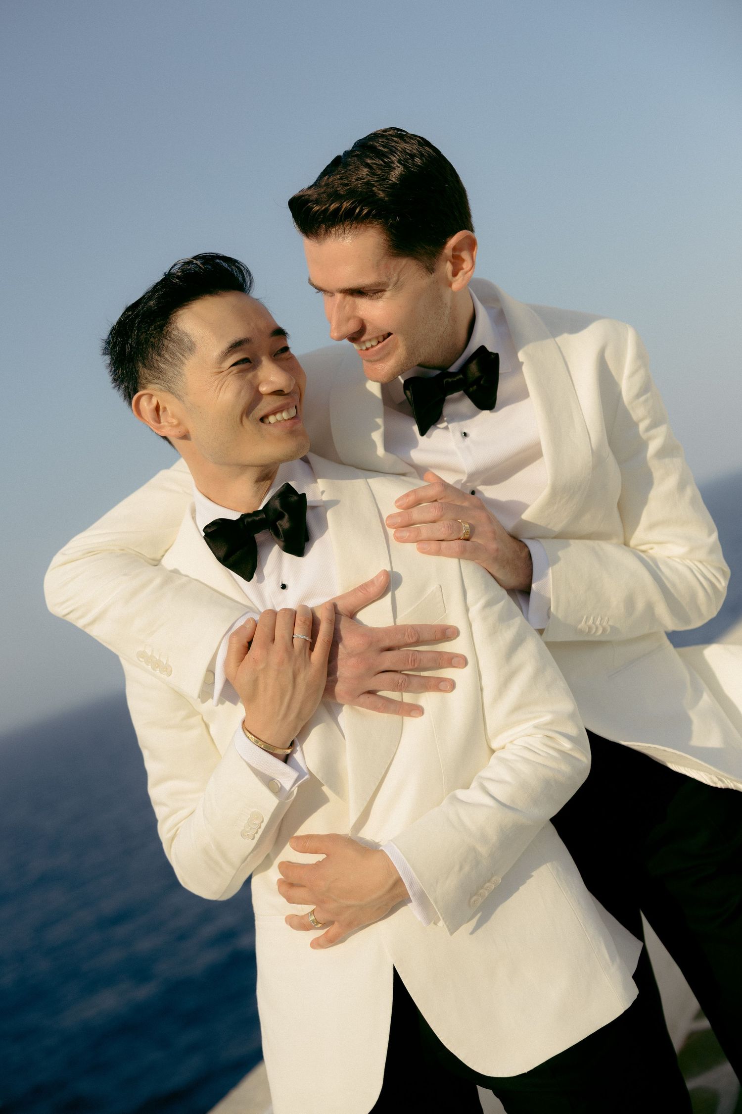 Two grooms in white tuxedos embrace and smile on a cliffside overlooking the ocean on their wedding day.