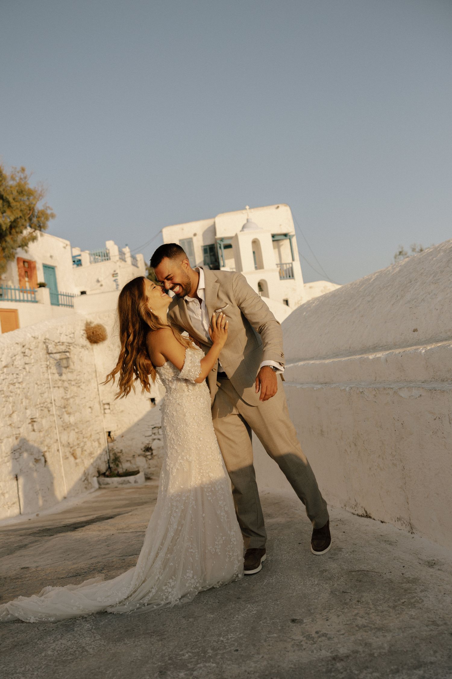A romantic wedding couple embrace in a dramatic dip pose against white buildings in Santorini at sunset.