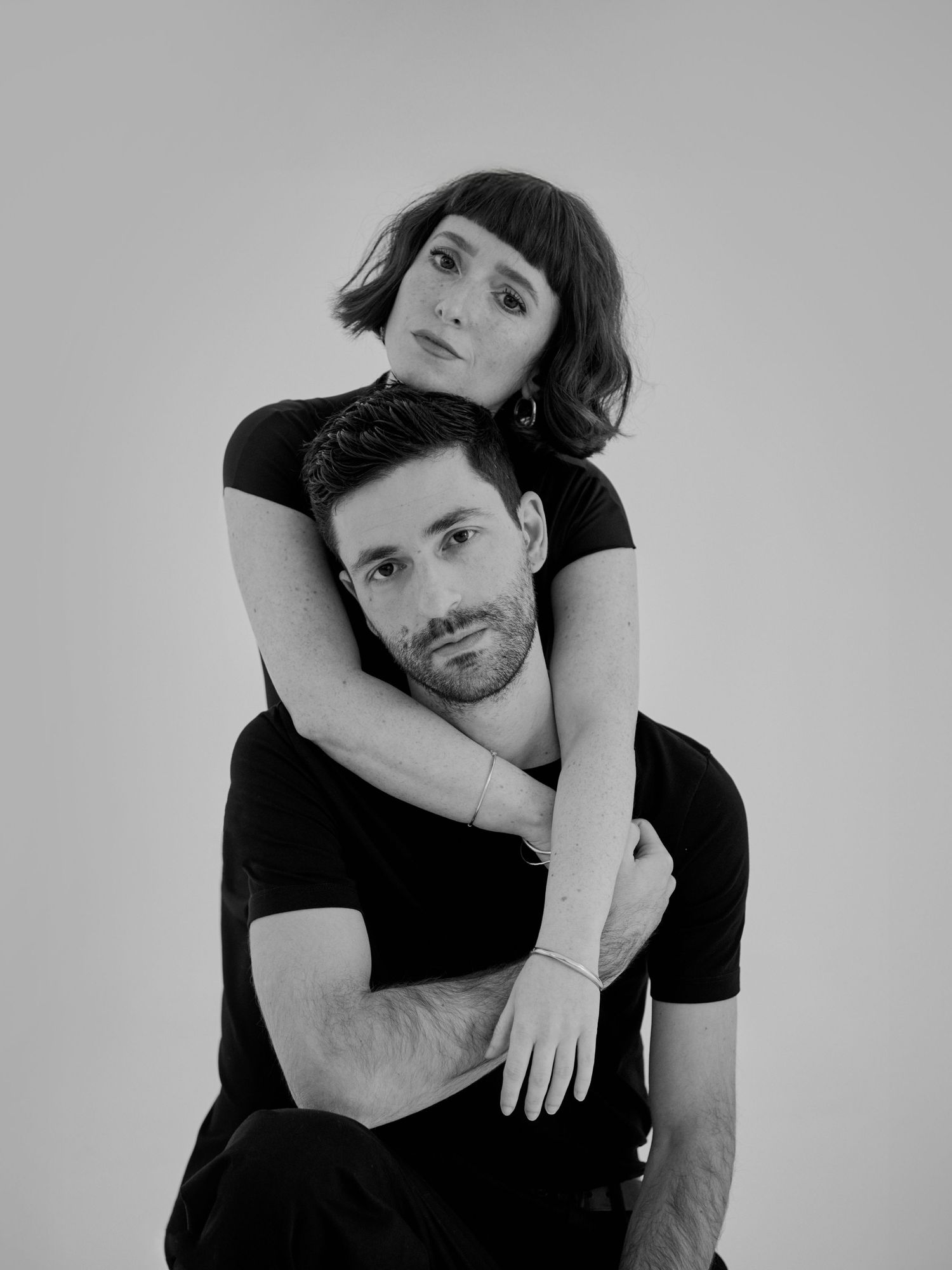 Black and white portrait of a couple, woman embracing man from behind, both wearing black tops.