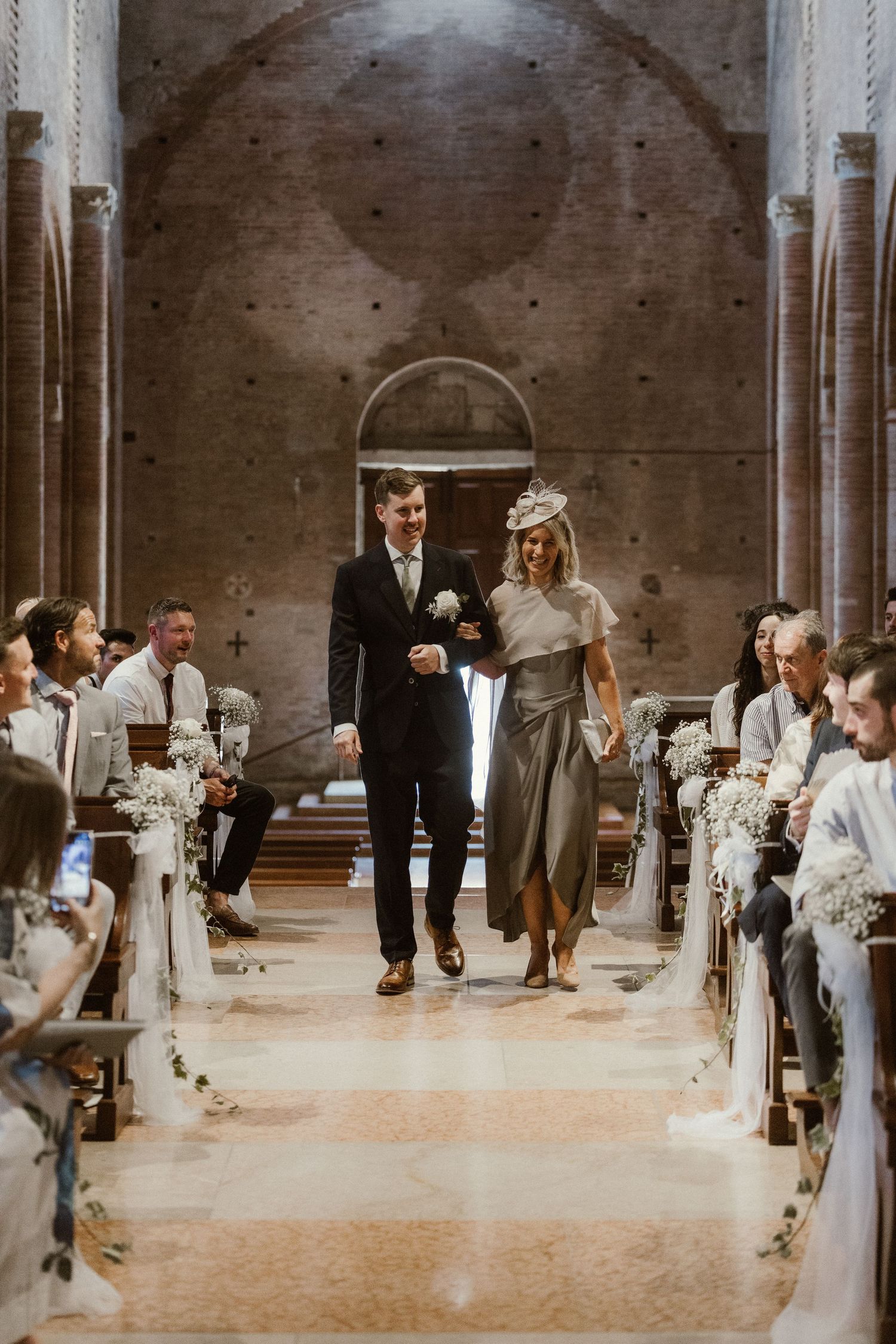 Bride and groom walk down the aisle of a stone church, surrounded by guests and white floral decorations.