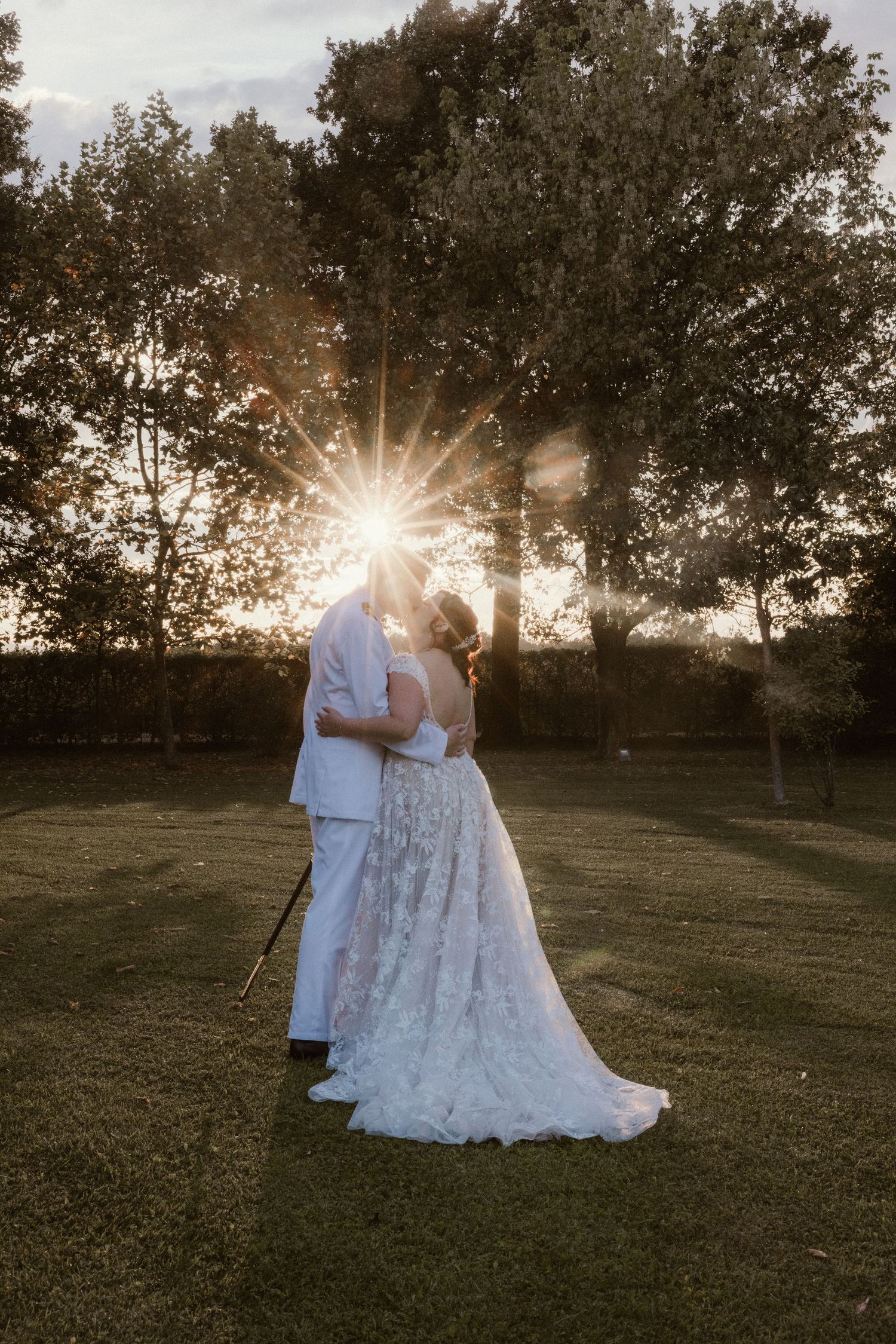 Bride and groom kissing at sunset with golden sun rays bursting between them in a lush green garden.