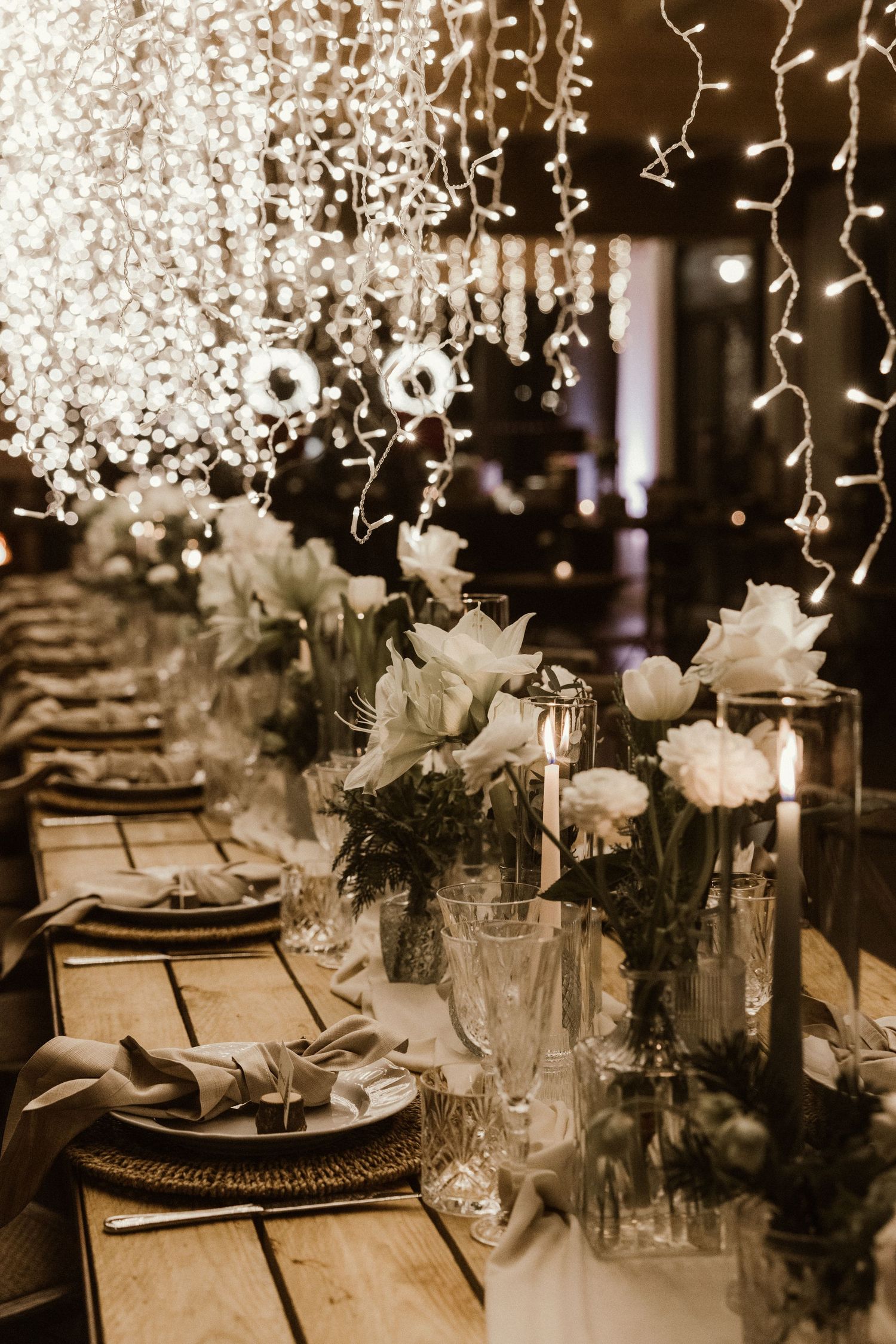 Elegant wedding reception table with white floral centerpieces, candles, fairy lights, and woven charger plates in warm lighting.