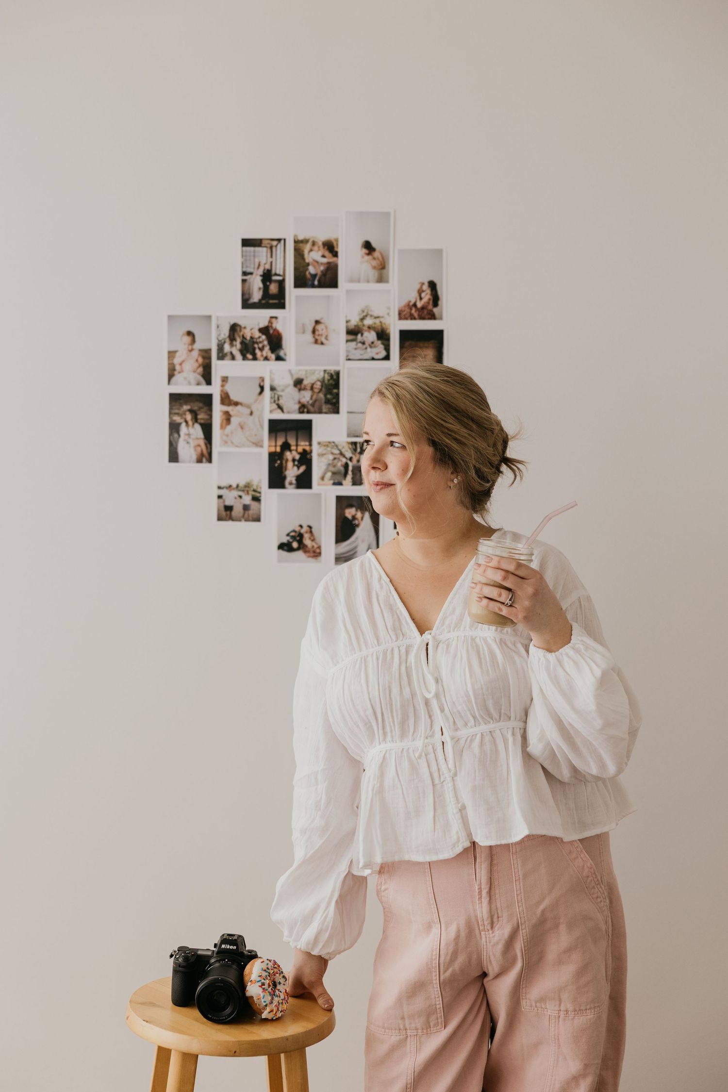 Photographer in a white blouse and pink pants stands thoughtfully in a minimalist studio with photos on the wall.