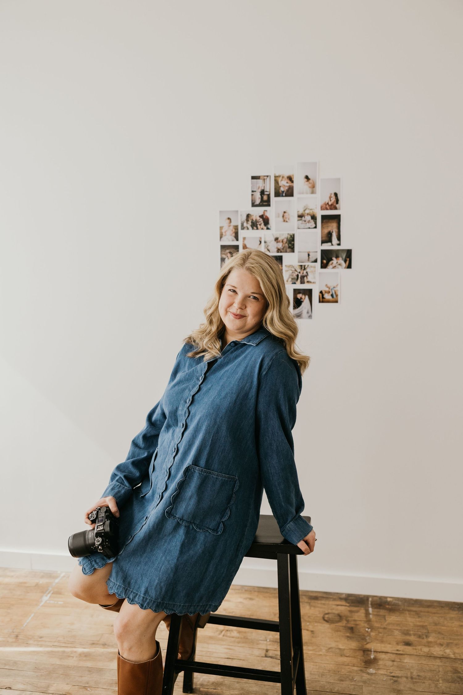 Woman in blue denim dress sits on stool against white wall with vintage photos displayed behind her.