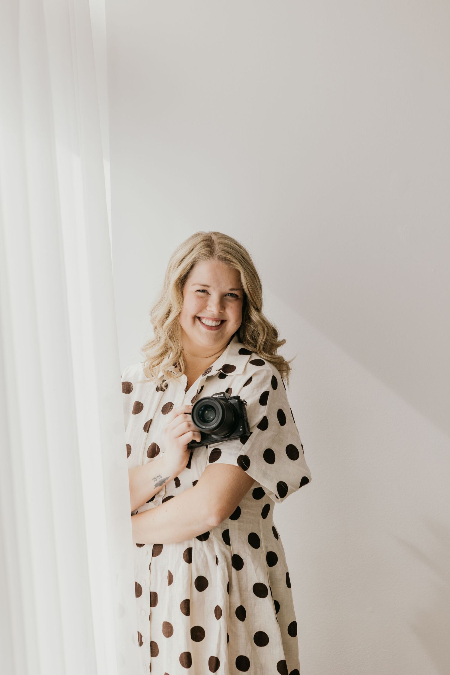 A person wearing a white polka dot dress holds and cuddles with a small black and white cat against a light background.