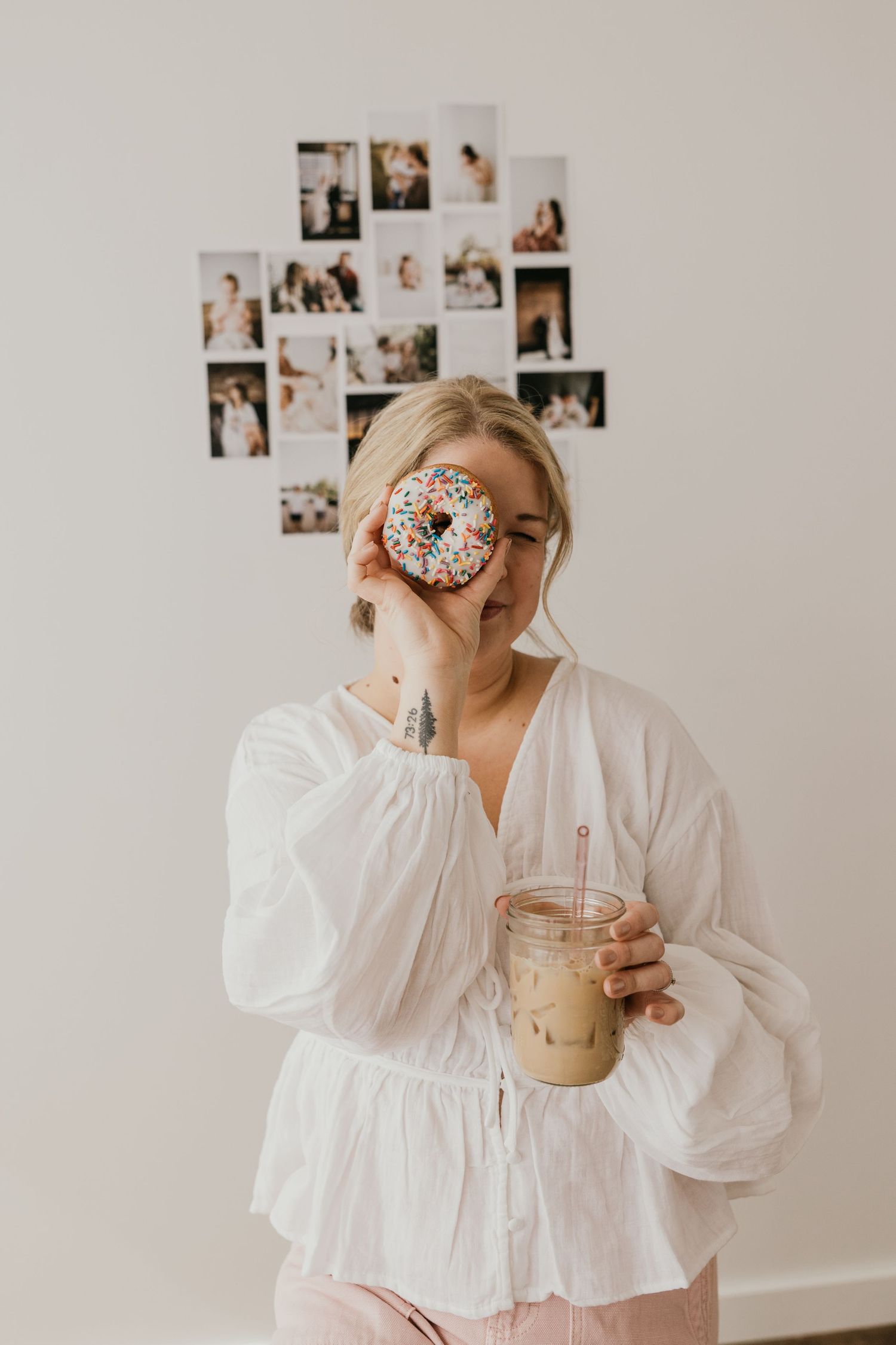 Woman in flowy white dress poses playfully with donuts and coffee against white wall with photos.