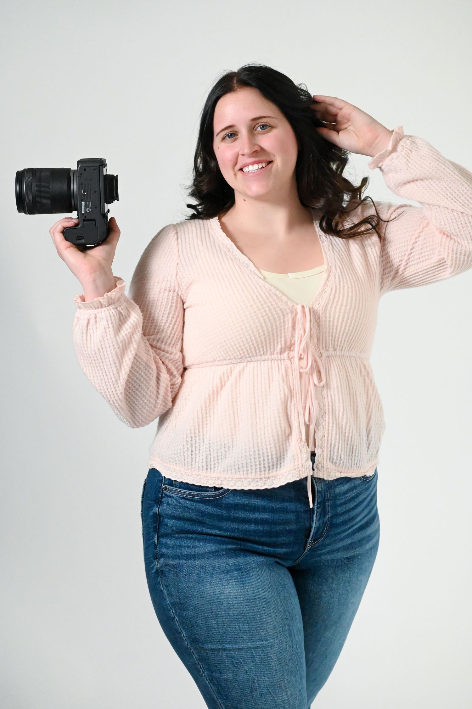Smiling woman holding a DSLR camera, wearing a pink blouse and jeans, posing against a white background.