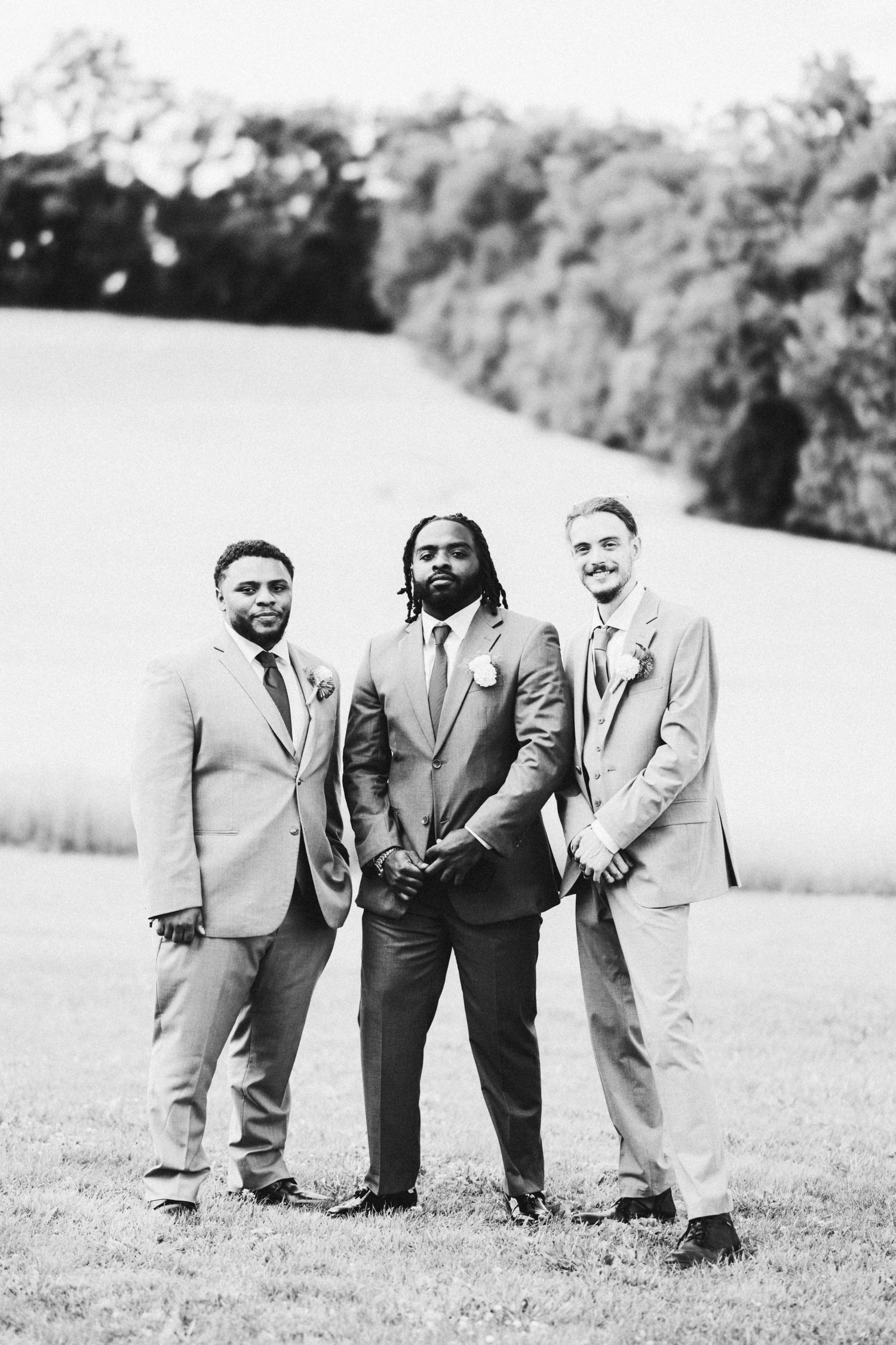 Black and white photo of three groomsmen in suits posing outdoors on a grassy field with trees in the background.