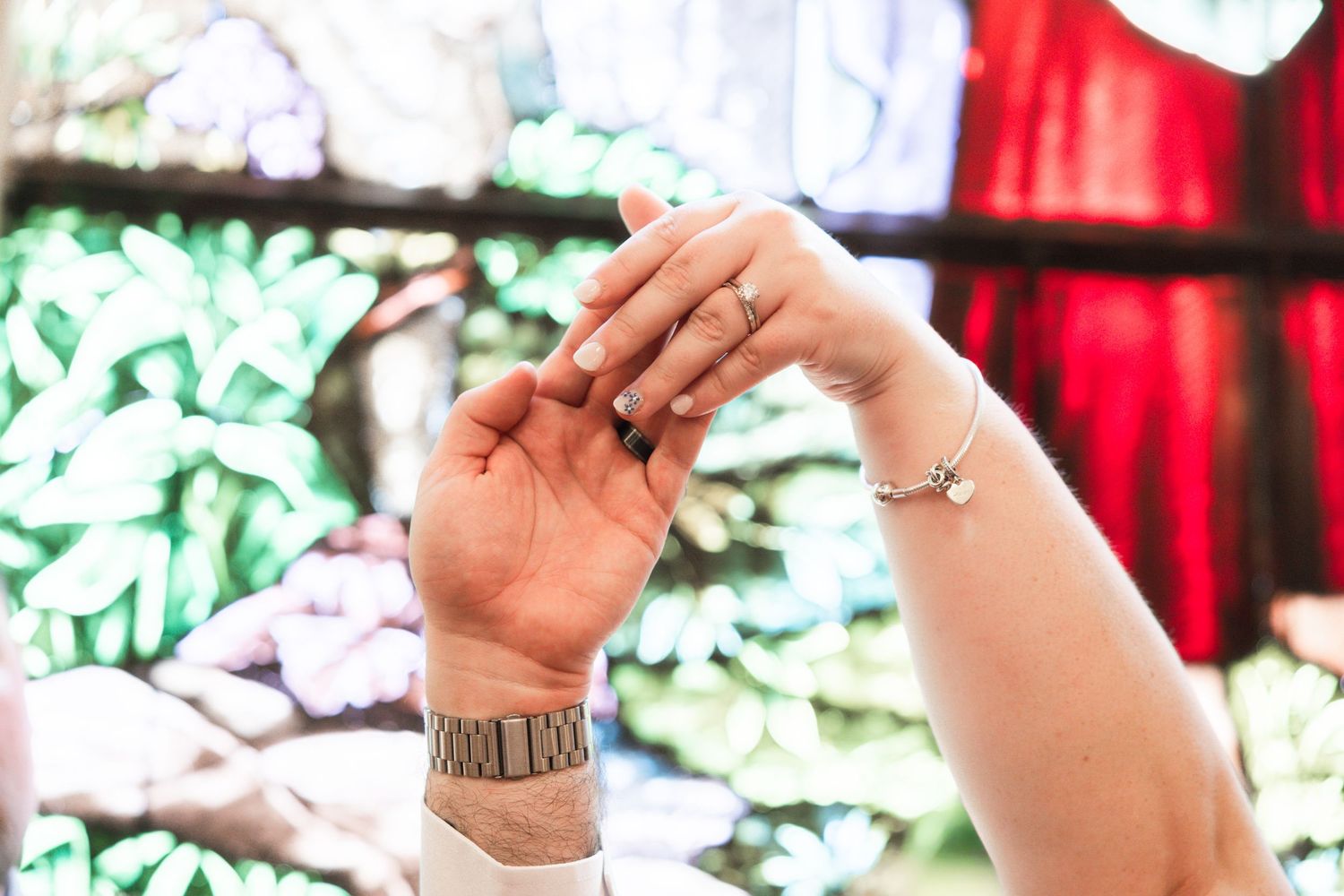 Two hands touching with a Roman numeral wrist tattoo and bracelet, set against colorful stained glass background.