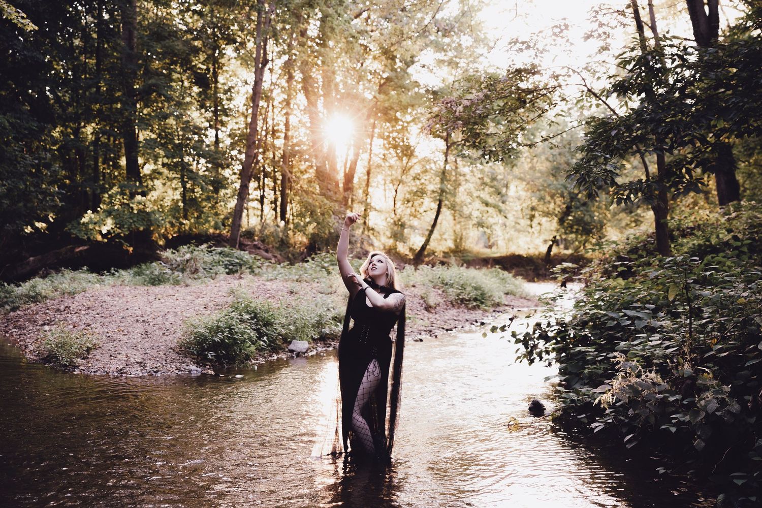 Woman in black dress standing in a sunlit forest creek with arms raised toward the sky.