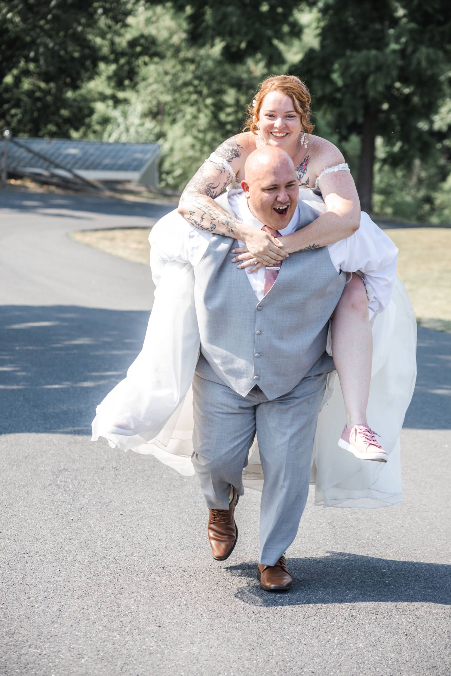 Laughing groom in gray suit gives bride in white wedding dress a piggyback ride outdoors.