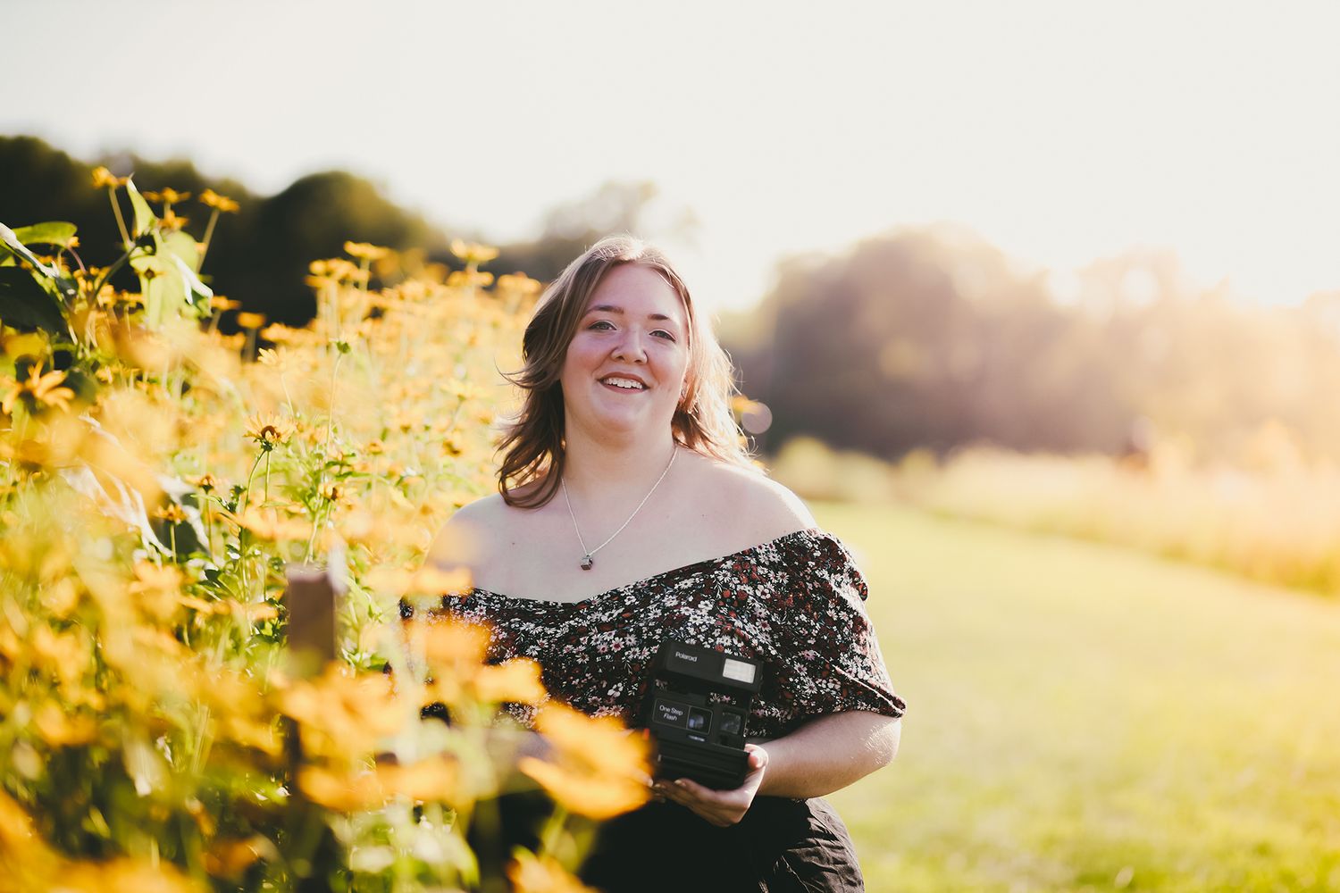 Smiling woman holding a camera in a sunlit field of yellow flowers, wearing a black off-shoulder dress.