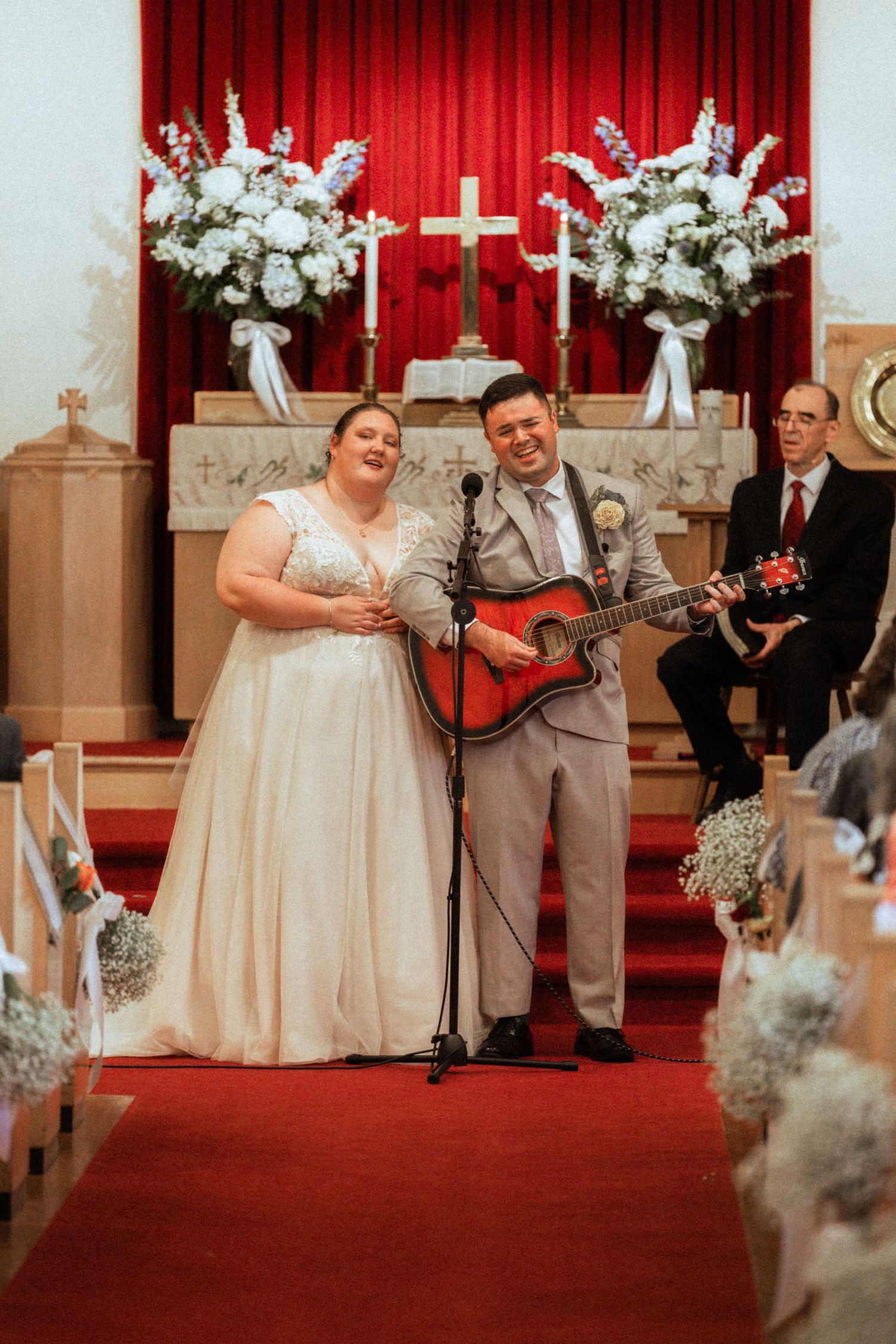 Bride and groom singing at church altar, groom playing red acoustic guitar, white floral arrangements and cross backdrop.