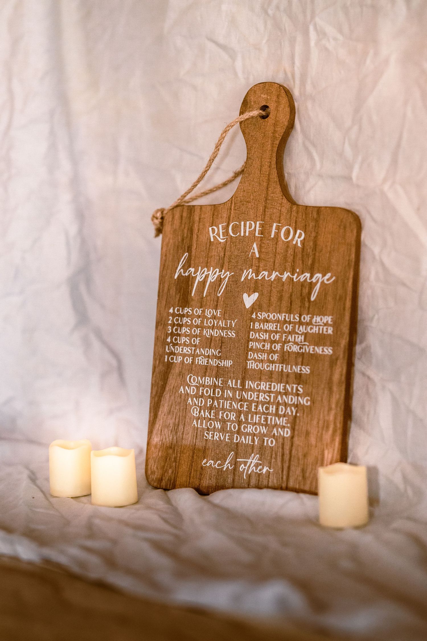 Wooden cutting board with Recipe for a Happy Marriage engraved, surrounded by flameless candles on white fabric.