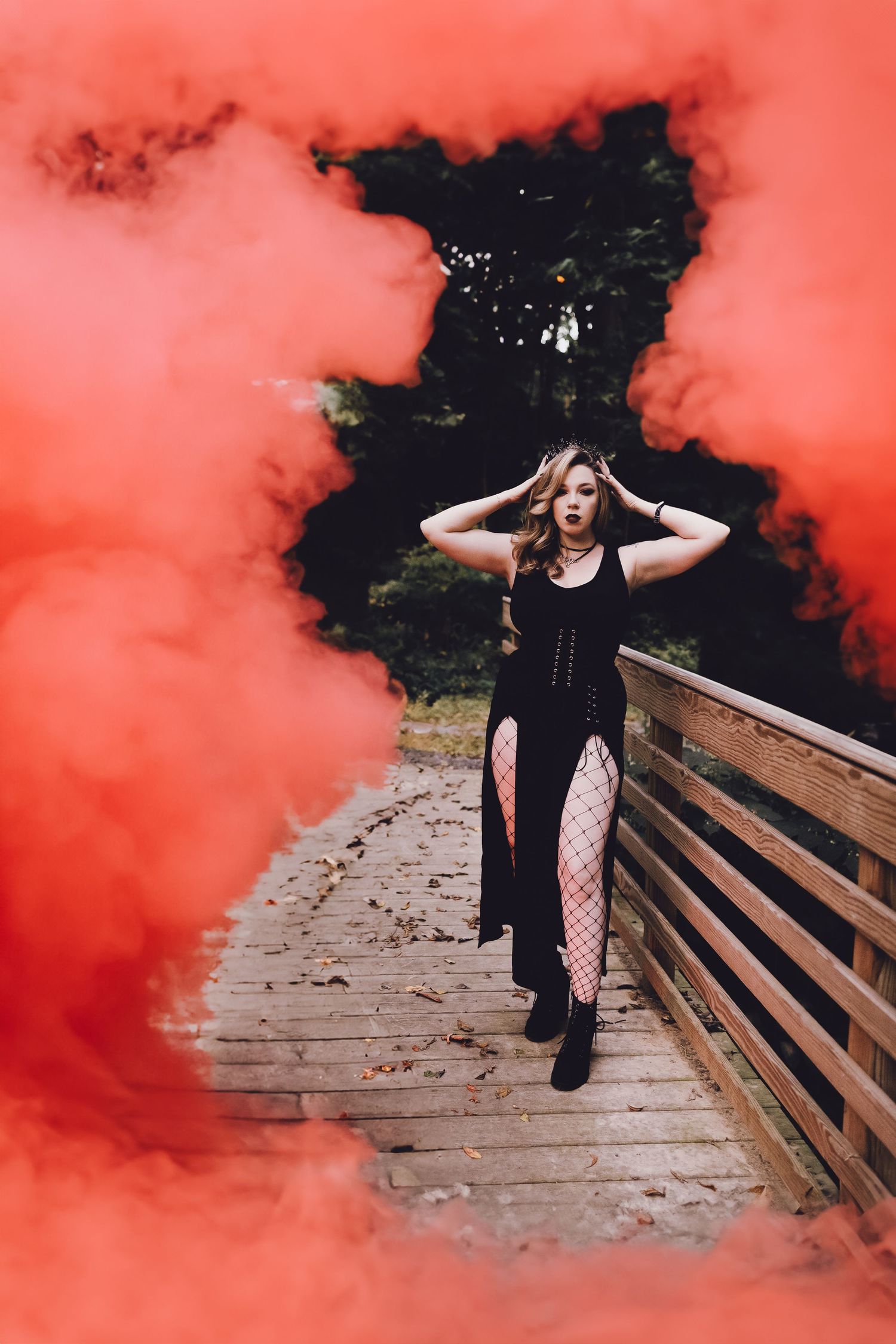 Woman in black corset dress surrounded by dramatic red smoke on a wooden bridge outdoors.